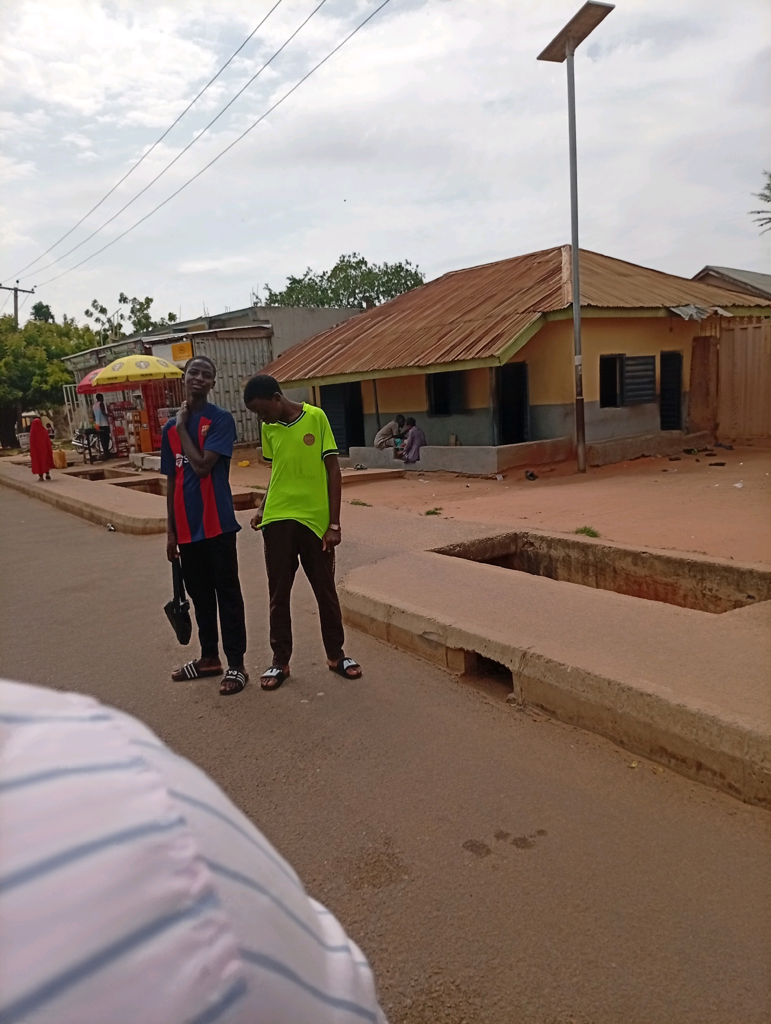 The image depicts a street scene in Garko, Nigeria. Two young men are standing on the paved road, facing the camera. The man on the left wears a blue and red striped t-shirt and black pants, carrying a black umbrella in his right hand. The man on the right wears a neon green t-shirt and dark pants. Behind them, a small, yellow building with a corrugated metal roof is visible. A solar-powered street light stands tall to the right of the building. Further in the background, a red-clad figure is walking away from the camera, and a yellow umbrella shades a small market stall. The ground is a mix of asphalt and reddish-brown soil, with a concrete drainage channel running along the side of the road. The sky is overcast with scattered clouds.
