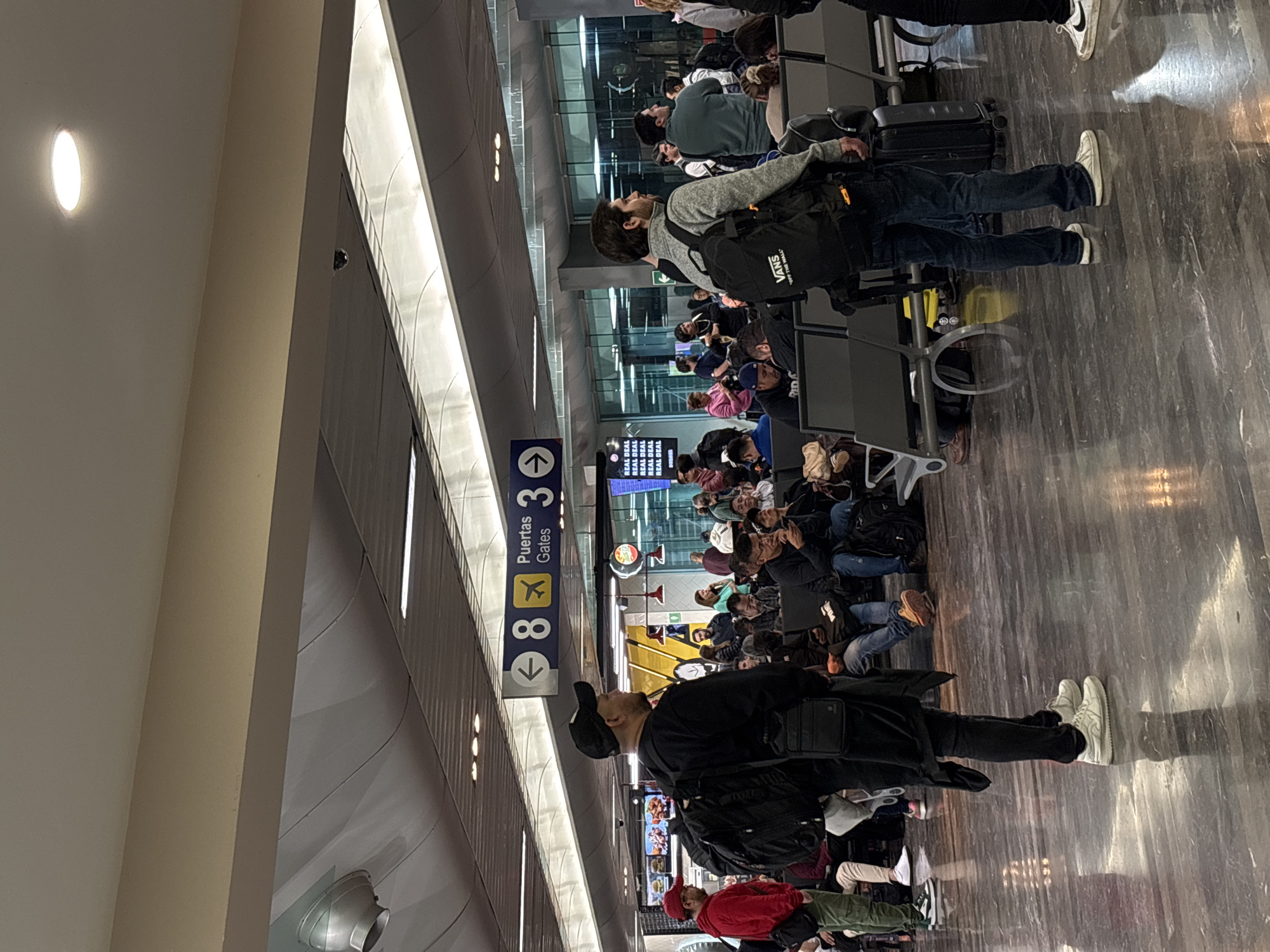 The image shows a waiting area, likely an airport gate, crowded with seated individuals and some standing. Most are facing the direction of the gate sign, which reads "Gates 3-8". People are dressed in a variety of casual attire. Some have luggage nearby. Overhead are ceiling lights and signage. The scene suggests a moment before boarding a flight.