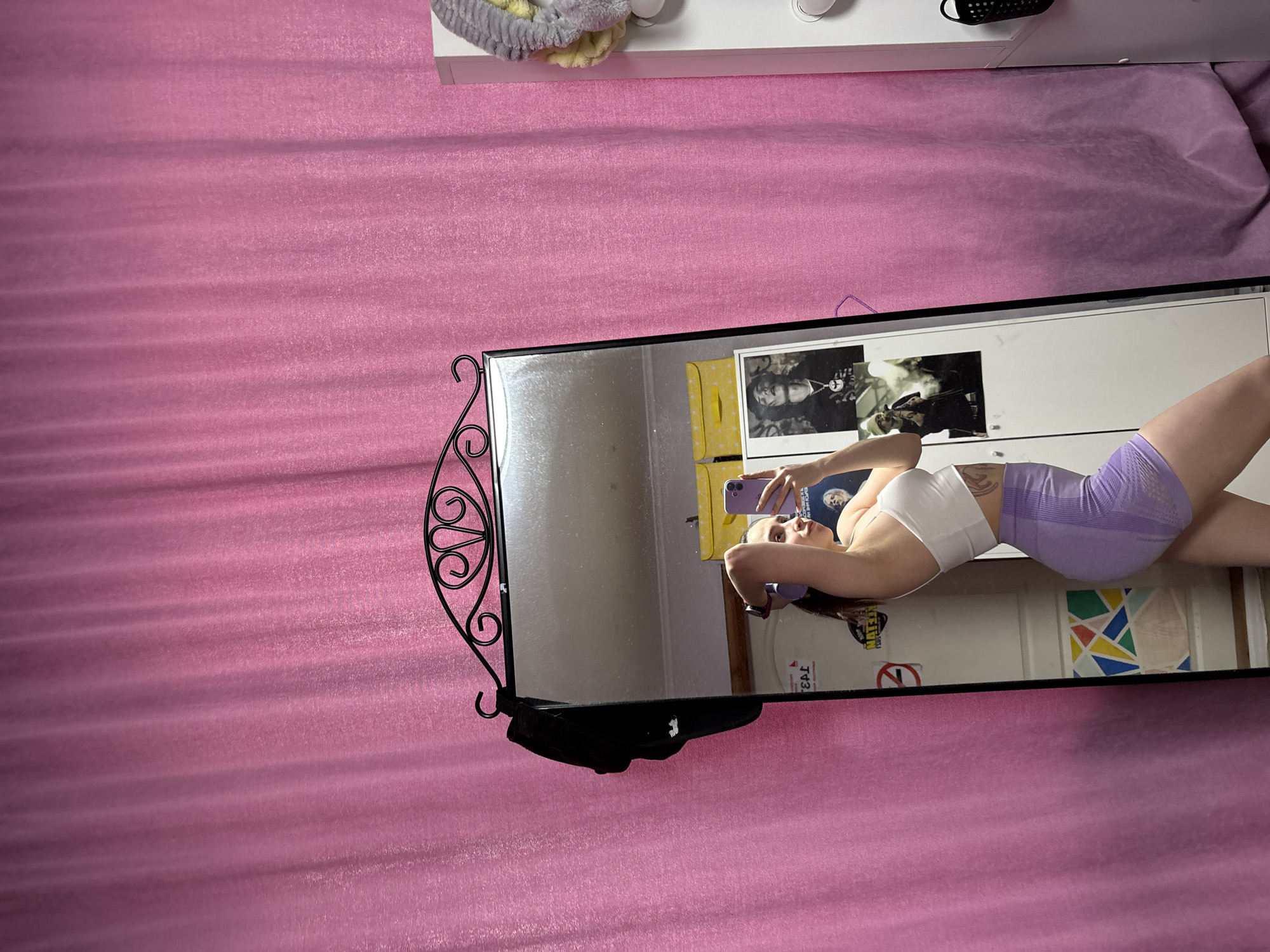 A person is visible in a mirror's reflection; they are fair-skinned and wearing a white sports bra and light purple shorts. The person is holding a purple cell phone, seemingly taking a photo of themselves. The background reflected in the mirror includes a white wall, posters, and possibly the edge of furniture. The mirror itself is ornate with black metal designs and is hanging on a pink textured wall. The location in the city of Прикамский, Russia is not evident.