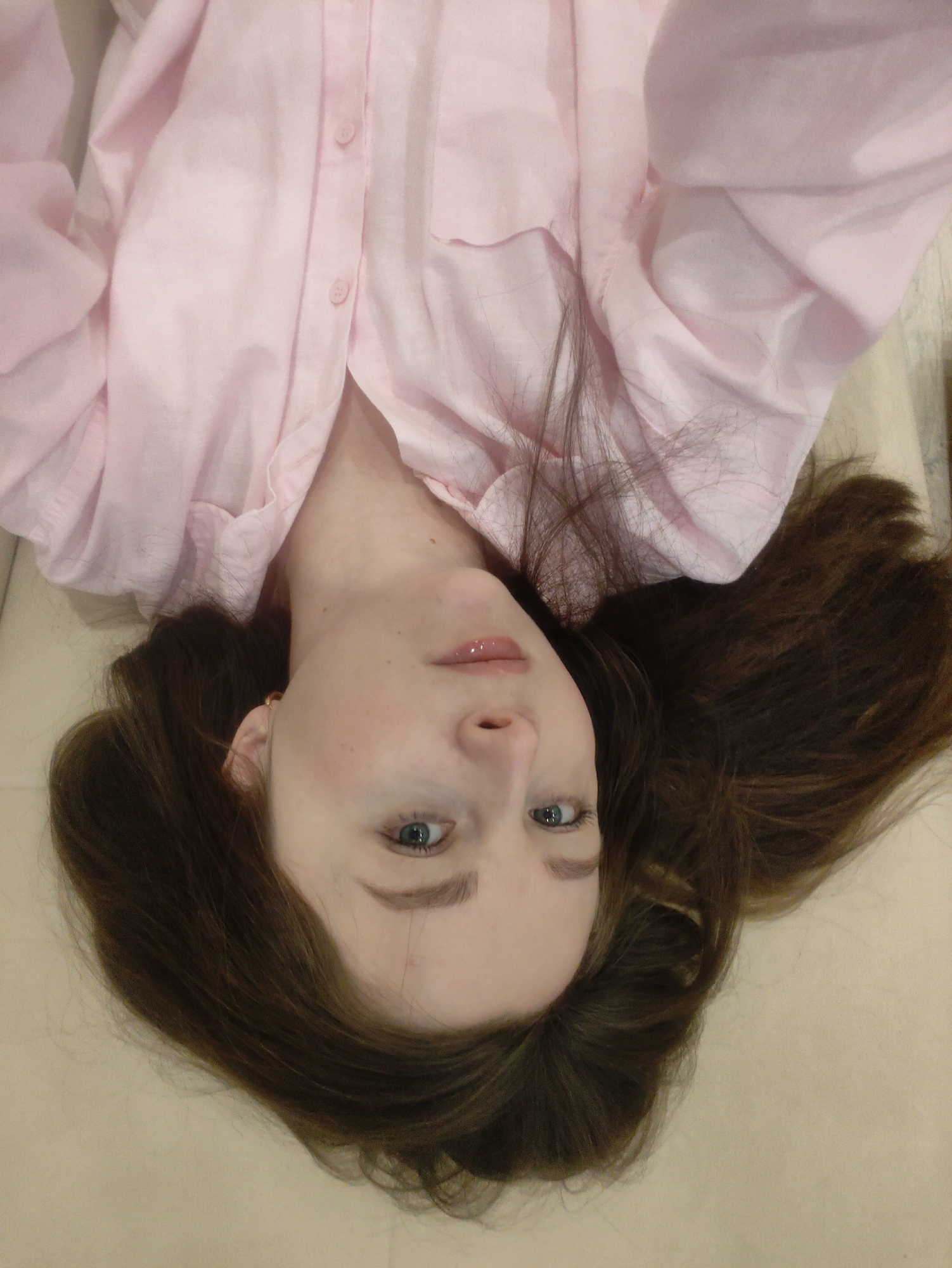 A woman with long, brown hair and blue eyes is lying down, facing upwards. She is wearing a light pink button-down shirt. The background appears to be a light-colored surface, possibly a floor or bed. The location is unknown.