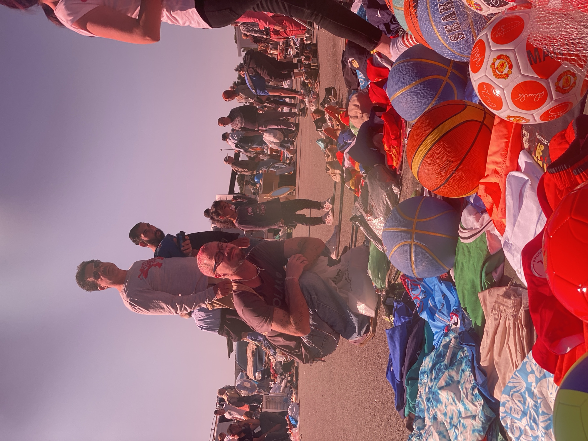 The image captures an outdoor market scene. People are browsing through various items for sale, including clothes and sports balls. The market is situated on a paved road. In the foreground, there are piles of clothing and a collection of basketballs and soccer balls. A man is crouched among the items. In the background, more people are milling around different stalls. The location appears to be in Podgorica, Montenegro.