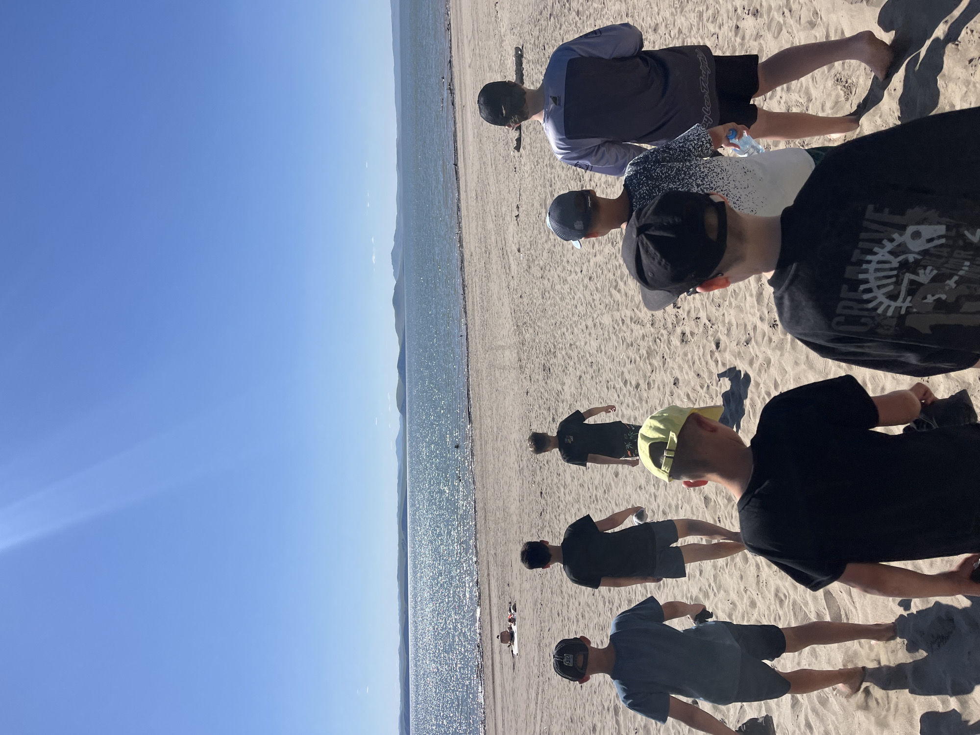The image shows a group of young males walking on a sandy beach towards a body of water under a clear blue sky. The sun is bright, casting shadows on the sand. In the distance, there is a wide body of water with a shimmering surface, and beyond that, a range of hills or mountains. The beach appears to be a natural environment, with some driftwood visible. Some individuals are wearing baseball caps. There is no visual information in the image to suggest this location is in the city of Krasnoturansk, Russia.