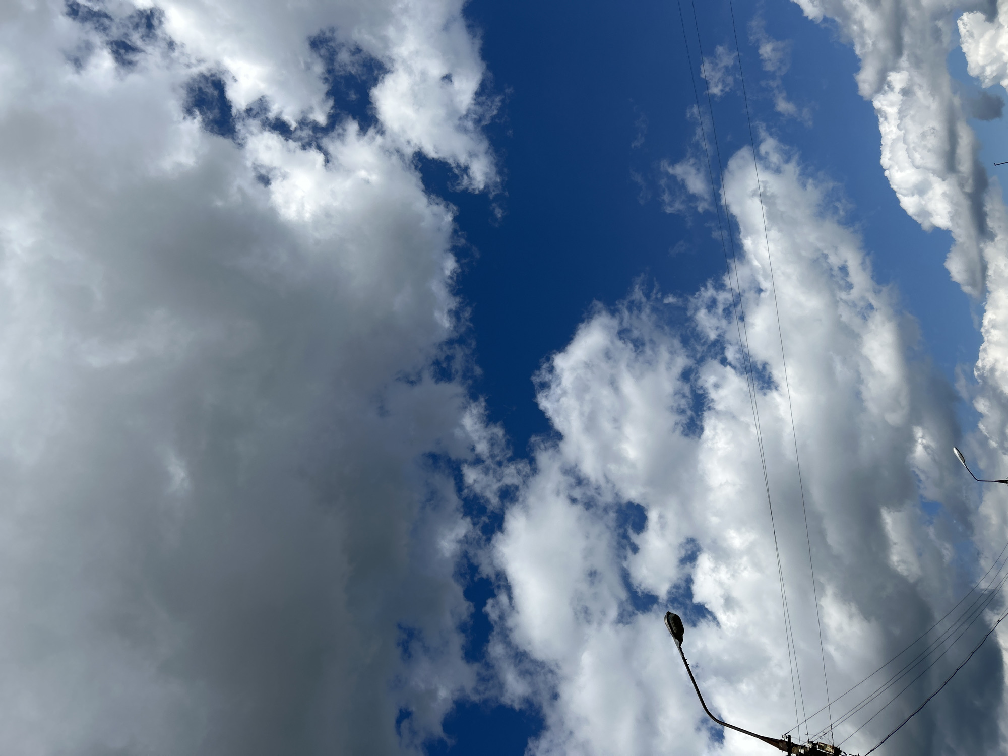 The image captures a view of the sky with clouds and utility poles, seen from street level in Zelenograd, Russia. A dark, curved street lamp extends from the left side of the frame, supported by a pole with visible wires and electrical equipment. In the distance, another street lamp is visible, smaller in the frame. The sky is a deep blue, partially obscured by a mix of large, dark, and white cumulus clouds. Several thin wires are strung horizontally across the upper portion of the image. The scene appears to be set during daylight hours.