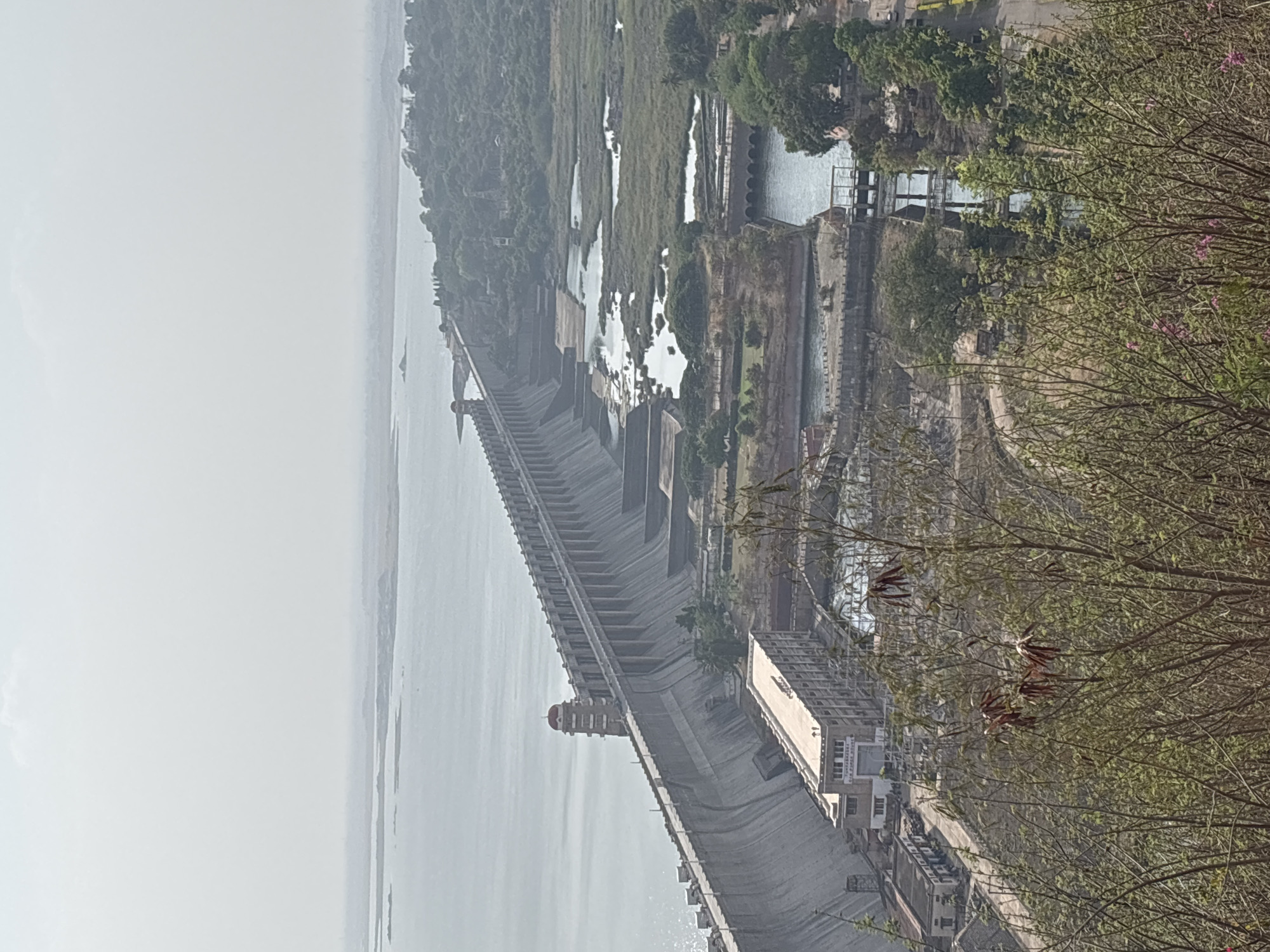This image depicts a dam in Hosapete, India, taken from an elevated position. The dam stretches across the frame, with a large body of water on one side and what appears to be a reservoir or river on the other. The structure is made of concrete with a series of stepped levels. There is a small tower or building located along the top edge of the dam, slightly left of center. Buildings sit to the side of the dam. The foreground is filled with tree branches, obscuring part of the view. The day seems overcast or hazy.