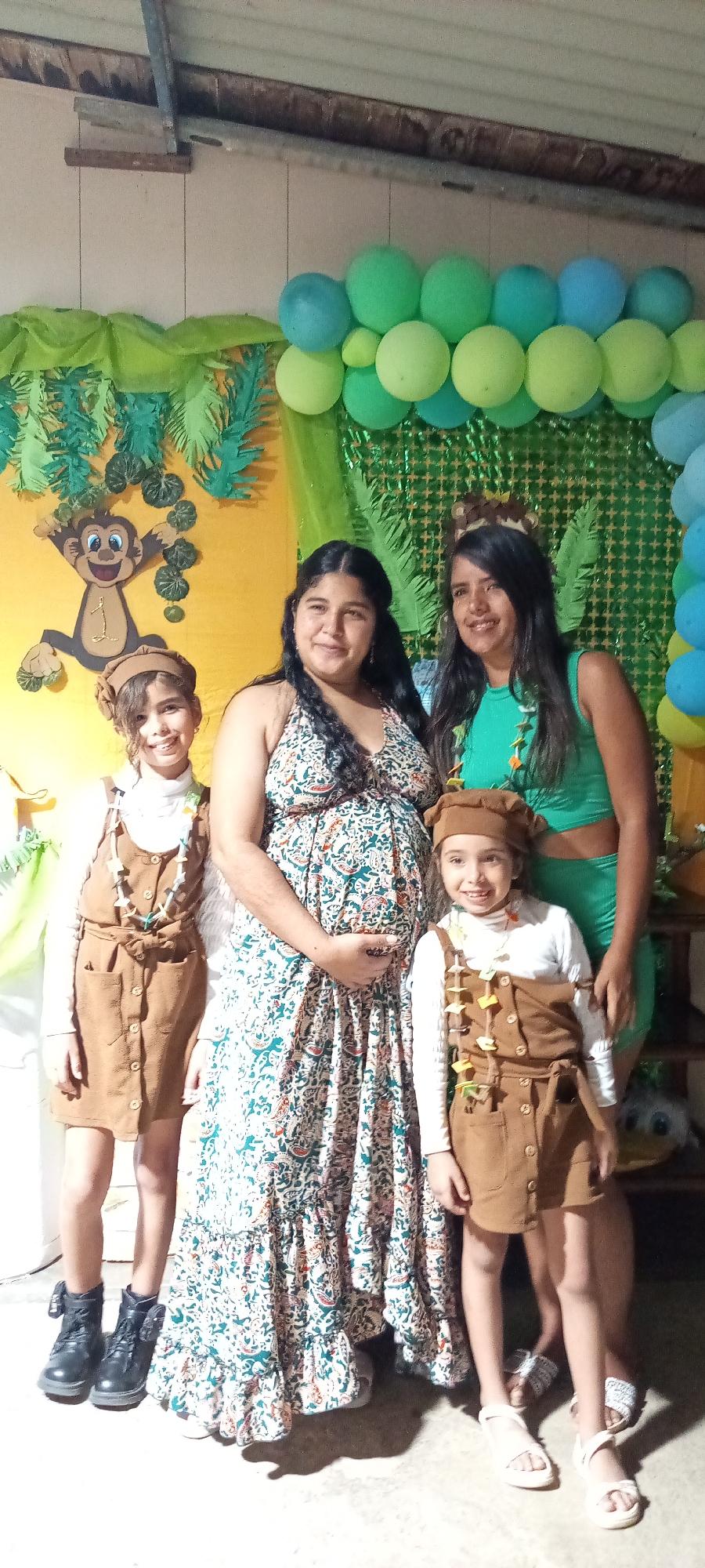 The image shows a group of four individuals posing in what appears to be a decorated indoor setting for a party or celebration, possibly in the city of Unknown, Unknown. The background features a yellow wall with a cartoon monkey graphic, along with green leafy decorations and a balloon garland in shades of green and blue.
From left to right:
A young girl stands wearing a brown dress-like garment over a white long-sleeved shirt, a brown hat, a necklace, and black boots.
A woman stands next to her, wearing a long, patterned dress. Her left hand is resting on her pregnant belly.
Another woman stands next to her, wearing a green two-piece outfit.
To her right, another young girl, dressed similarly to the first girl in a brown outfit, white shirt, brown hat, a necklace, and white sandals.
The children are smiling and seem to be part of the celebration. The lighting is soft, and there is evidence of an indoor ceiling in the background. Overall, the atmosphere seems celebratory.