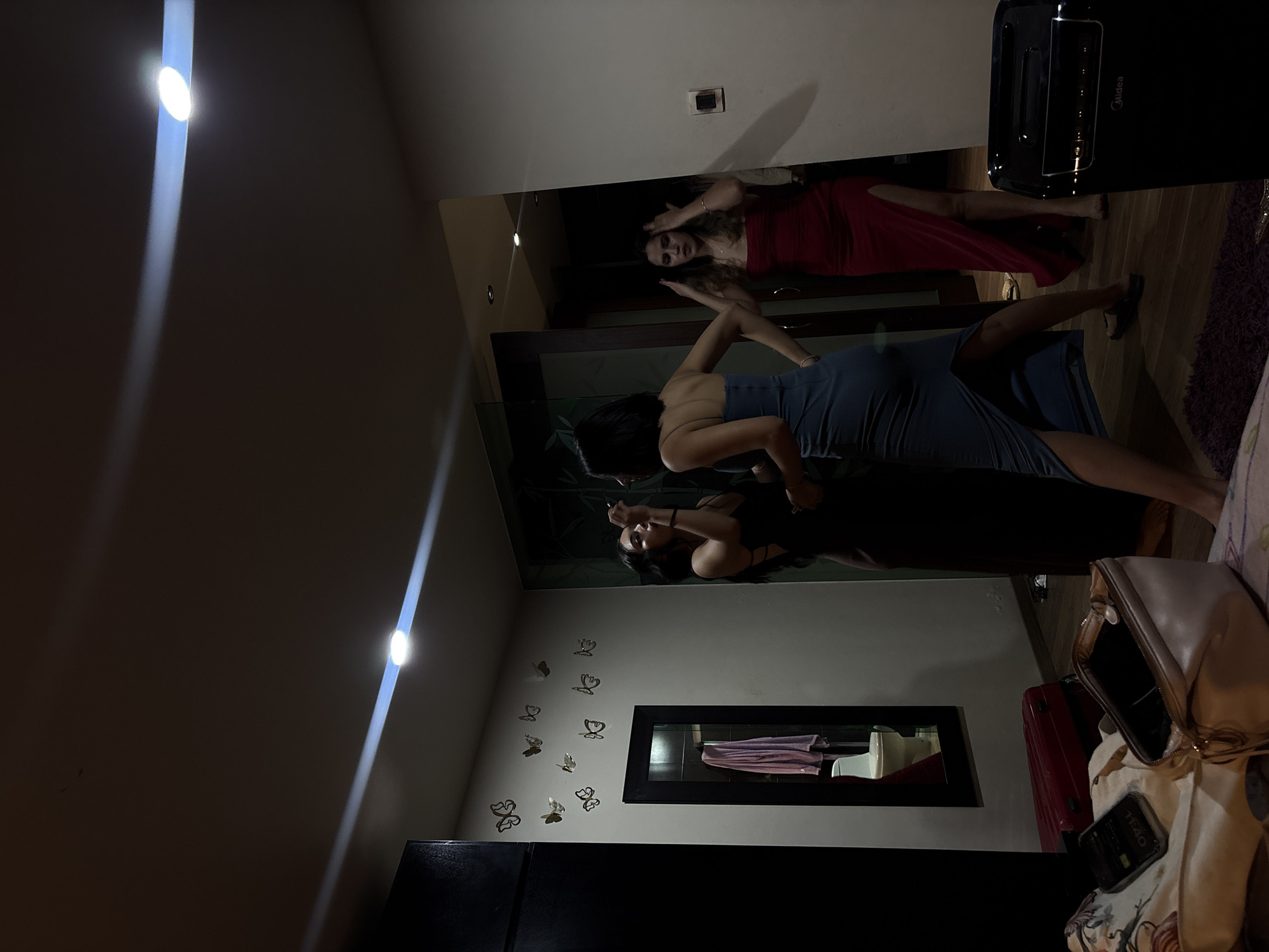 In a dimly lit room, three young women are present. One, wearing a dark dress, is applying makeup to another woman wearing a blue dress. The third woman, in a red dress, stands near a doorway, her hands on her head. The room contains a mirror reflecting a toilet and a pink towel, a black wardrobe, decorative butterflies on the wall, a red suitcase, and an open makeup bag with a phone on top of some fabric. A black appliance with the brand "Midea" is visible in the foreground. The setting appears to be a private residence, potentially a hotel room, in San Andrés Cholula, Mexico, indicated by the presence of the Midea appliance which is commonly found in Mexican homes.
