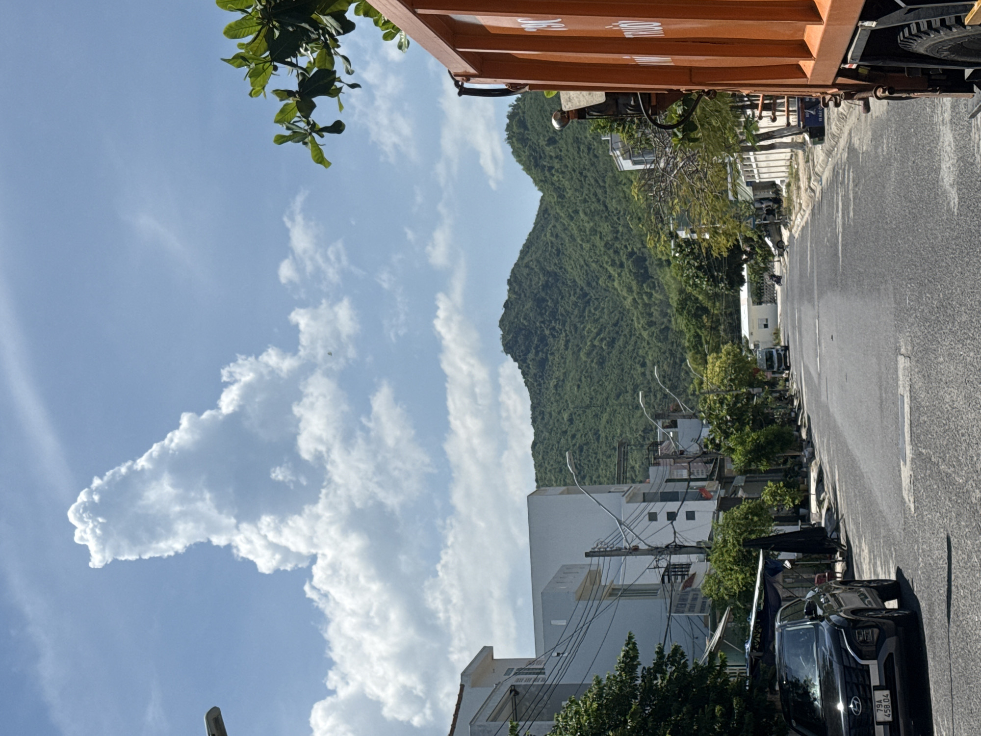 This image depicts a street scene in Bac Nha Trang Ward, Vietnam, on a bright, cloudy day. A narrow, paved road runs through the center of the frame, flanked by low-rise buildings and lush green vegetation. In the foreground, an orange truck occupies the right side of the image, with its side prominently visible. On the left, a black SUV is parked, and further down the street, a white minivan can be seen. The background is dominated by a verdant, tree-covered mountain range under a sky filled with dynamic cumulus clouds. The overall impression is one of a coastal town or a community situated near a natural landscape.