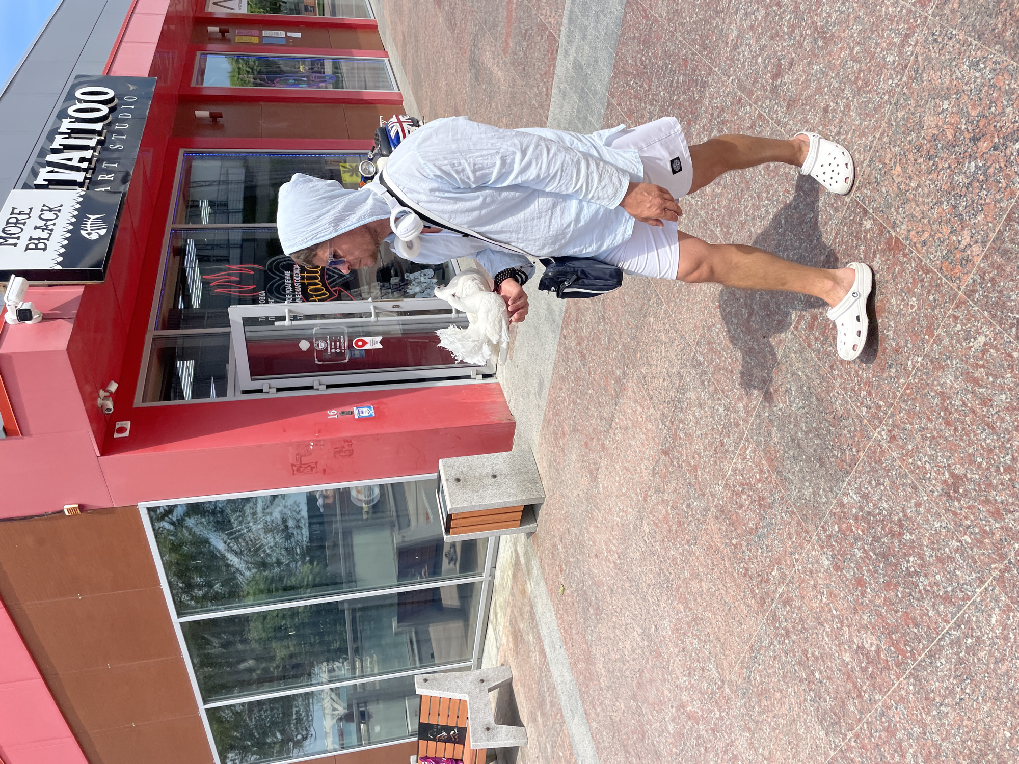 A man walks on a tiled sidewalk in front of a tattoo parlor. He is wearing a light blue hooded shirt, white shorts, and white Crocs. He has white headphones around his neck and a dark blue bag slung across his chest. In his left hand, he is holding two white doves. The tattoo parlor has a bright red exterior and a sign that reads "MORE BLACK TATTOO ART STUDIO". There are large windows with reflections of trees and a bench in front of the establishment. The ground is paved with red and gray tiles.