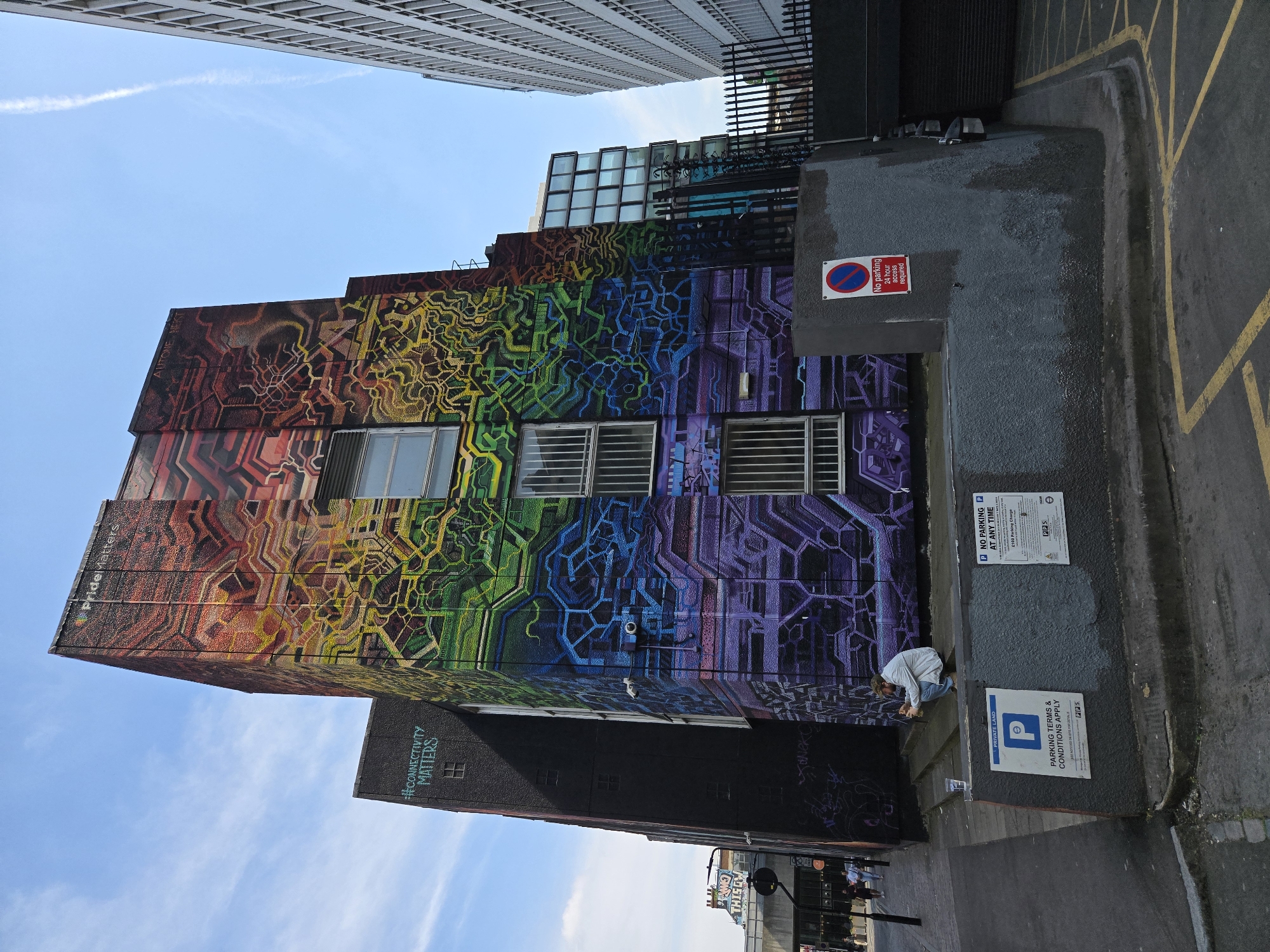 The image shows a building exterior in the city of London, United Kingdom, featuring a rainbow-colored mural spanning the facade. The mural, with a geometric design, transitions from red to violet across the building's height. Three windows with barred panes are visible within the mural. Adjacent to the building is a dark gray wall with posted signs, including "NO PARKING AT ANY TIME," "PARKING TERMS & CONDITIONS APPLY," and "No parking 24-hour access required." A person in a light-colored shirt is crouched near the wall. The building is flanked by taller buildings, one partially visible at the top and another at the bottom. The sky is visible with a few clouds.