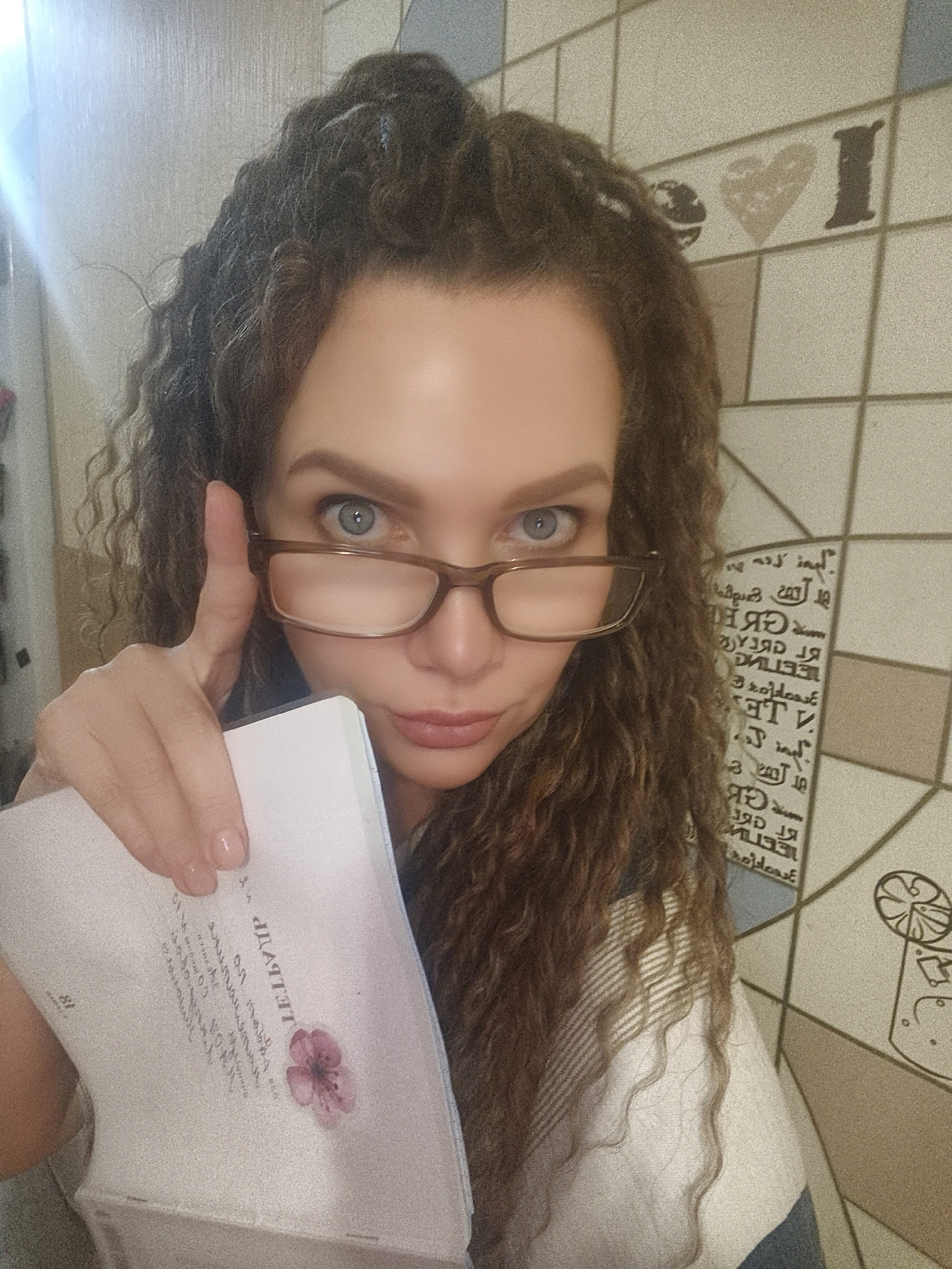 A woman with curly brown hair and wearing glasses is holding a book and pointing a finger towards the viewer. She has light skin and blue eyes. The book in her hand is white with some text and a pink flower graphic. The background consists of a tiled wall with various designs, including text and graphic icons. The location appears to be indoors, possibly a residence.