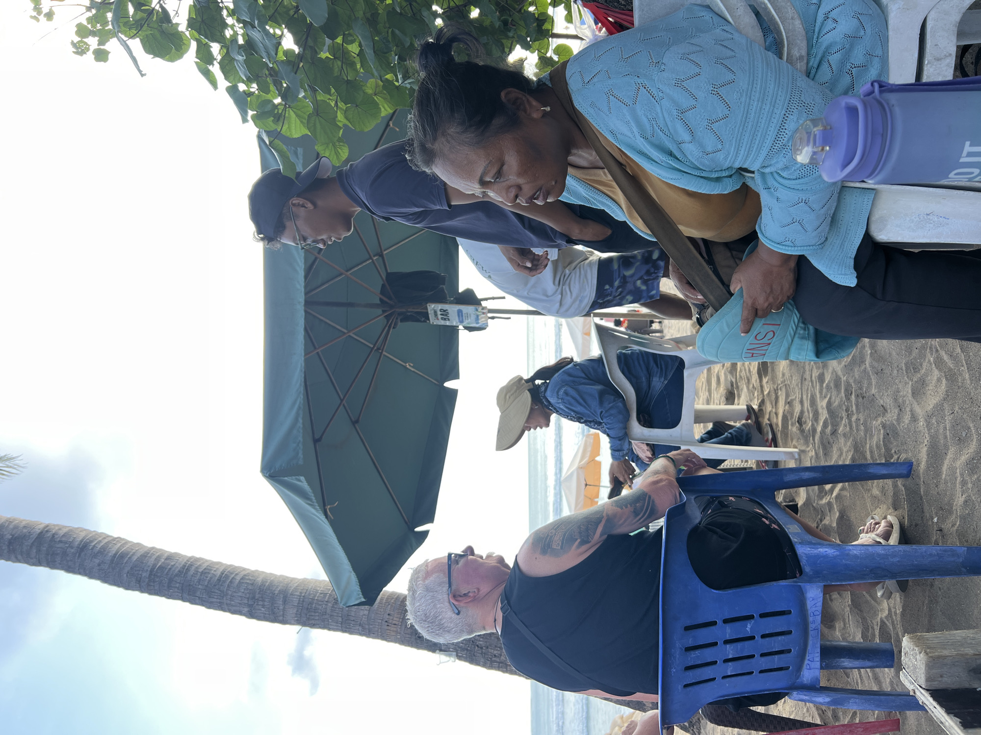 In Kuta, Indonesia, on a sandy beach, a group of people are seated under a large green umbrella. A Caucasian man with tattoos on his left arm, wearing a black tank top and sunglasses, is seated in a blue plastic chair facing the ocean. To his right, a woman in a denim jacket and a wide-brimmed hat is seated in a white chair. Further to the right, a woman wearing a blue cardigan over a tan top is seated in a white chair, holding a light blue bag and looking to her left. Standing behind them is a young man wearing a dark blue t-shirt and a baseball cap, looking to his left. A portion of a palm tree is visible to the left of the frame, and the ocean and sky are in the background. Other beach umbrellas and chairs are scattered around the scene.