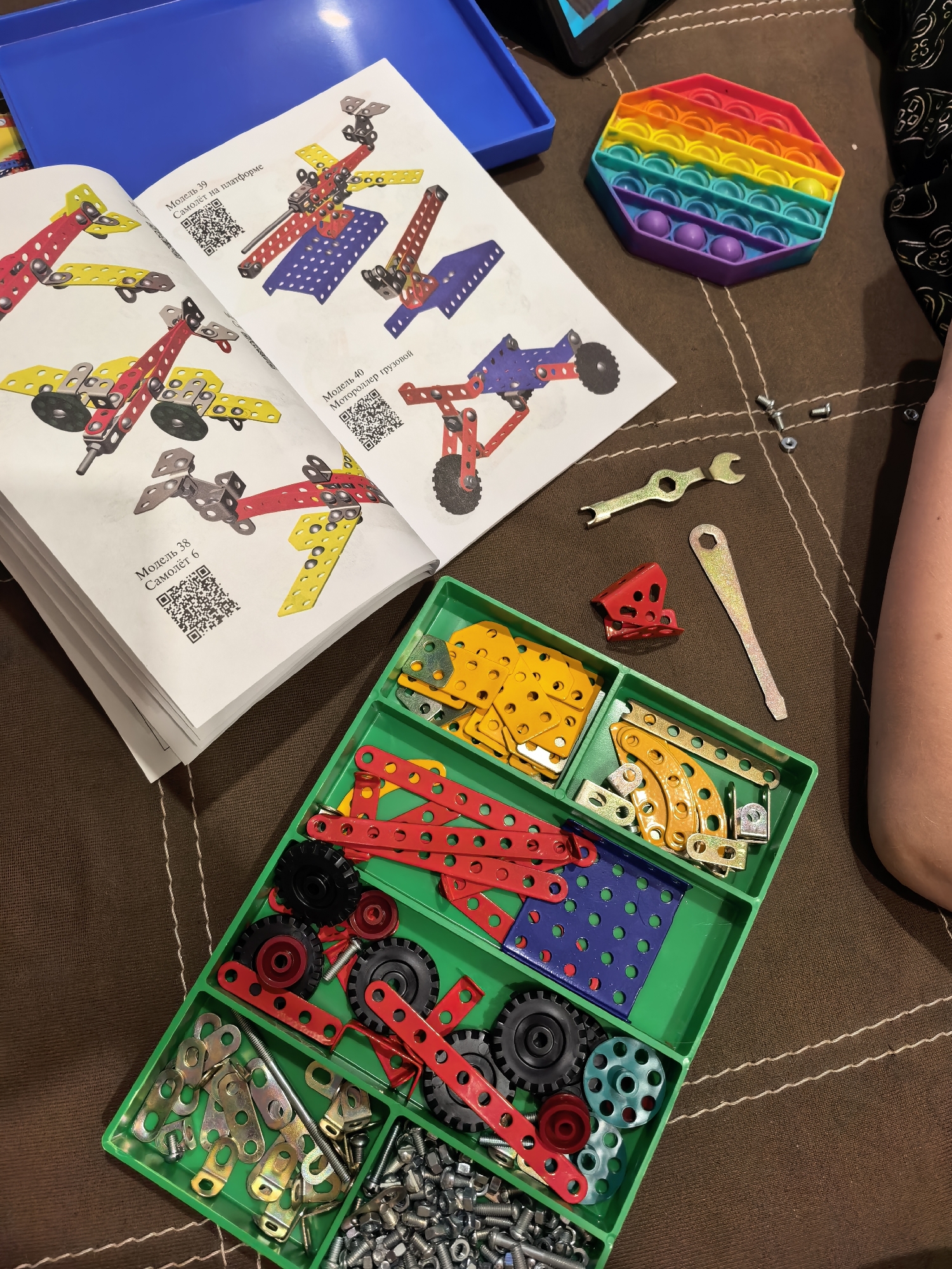 The image depicts a cluttered scene on a brown surface, likely a bed or table, with various objects scattered around. A green plastic container, segmented into compartments, holds an assortment of metal construction pieces in red, yellow, blue, and other colors, along with gears and fasteners like nuts and bolts. Next to the container, an open instruction manual displays diagrams of model airplanes and vehicles. A blue plastic tray sits nearby, and a colorful hexagonal pop-it fidget toy is also present. Tools, including wrenches, are placed on the surface. A person's arm is visible on the right side of the frame. The image does not provide evidence that the location is in Sterlitamak, Russia.