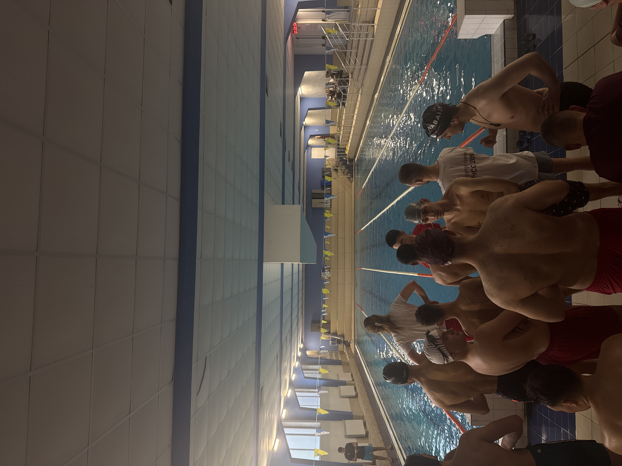 The image depicts an indoor swimming pool, seen from the perspective of someone standing at one end, looking down the length of the pool. The setting is a modern swimming complex with light-colored paneled walls and a blue stripe running along them, meeting a light-colored ceiling with embedded lighting.
In the foreground and extending along the near side of the pool, a group of approximately a dozen young men, mostly shirtless, are gathered. Many wear swimming caps and goggles, indicating they are swimmers. They appear to be waiting or preparing for an event. One swimmer in the foreground wears a black cap with "NABAIJI" written in white. Another individual in a white t-shirt, standing amongst the swimmers, has Cyrillic text visible, which appears to read "СПОРТЕН КЛУБ" (Sports Club) and "НСС 202" (NSS 202). Most of the individuals wear swim trunks, with some wearing red or patterned shorts.
The pool itself has several lanes marked by ropes, and further down the pool, starting blocks are visible, some bearing the brand name "Delfin." Yellow triangular flags hang above the lanes, typically used for backstroke turns. On a wall at the far end of the pool, a red digital timer or scoreboard displays "10:52:28," suggesting the time of day is late morning. The water in the pool appears clear and slightly disturbed, indicating recent activity or movement. The overall impression is that of a swimming practice session or a local competition in progress or about to begin at a sports facility in Sofia, Bulgaria.