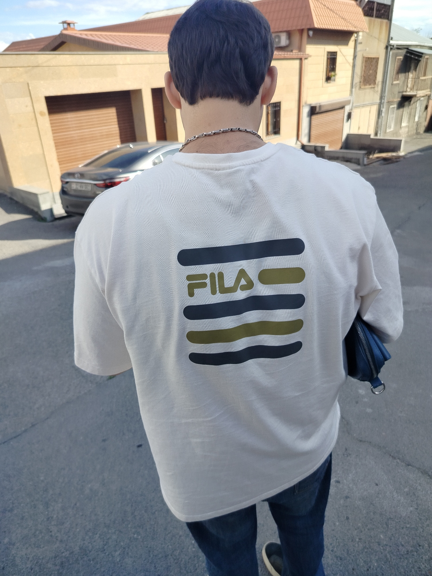 A man stands facing away from the camera on a street in Yerevan, Armenia. He wears a white t-shirt with a FILA logo consisting of horizontal lines in dark blue and olive green. He has a silver chain around his neck, blue jeans, and shoes with white soles. He carries a small, dark blue purse. Buildings with varied architecture stand in the background, including a garage with a closed brown door and a residential building with windows and a balcony. A silver car is parked in front of the garage. The ground is paved and appears to be part of a street or sidewalk. The sky is visible in the upper portion of the frame.
