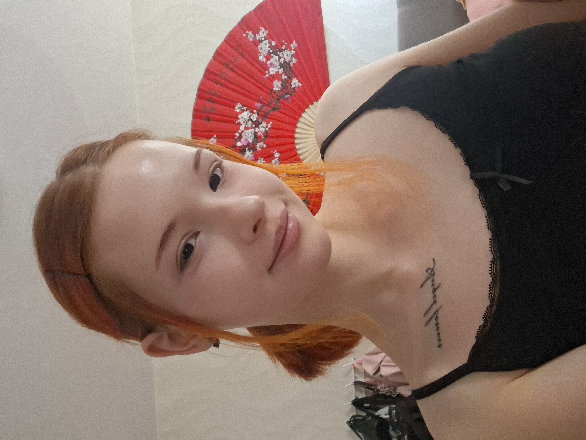 A close-up portrait of a young woman with red hair, fair skin, and dark eyes, is angled towards the viewer. She is wearing a black, lace-trimmed camisole with thin straps. A red folding fan, decorated with white floral designs, is positioned behind her head and shoulder. A tattoo is visible on her upper chest. The background appears to be a neutral-toned wall. The location in the city of Chaikovsky, Russia is undetermined.