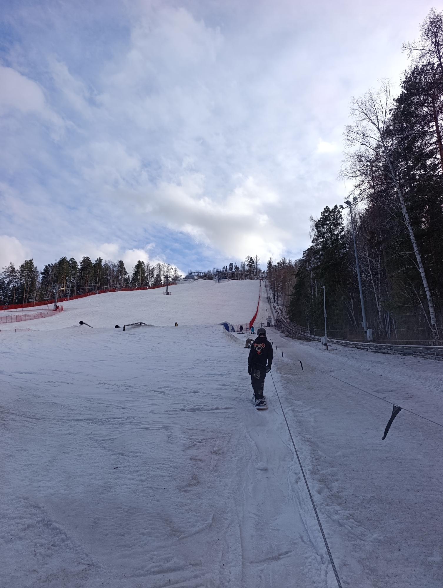 The image shows a snowy ski slope at the Bobrovy Log Funpark in Krasnoyarsk, Russia. A person wearing a black hoodie and riding a snowboard is being pulled uphill by a rope tow. The slope is marked with orange safety netting on the left side. Other skiers and snowboarders are visible in the distance on the slope. Trees are present on both sides of the slope, and the sky is cloudy. There are also several light poles.