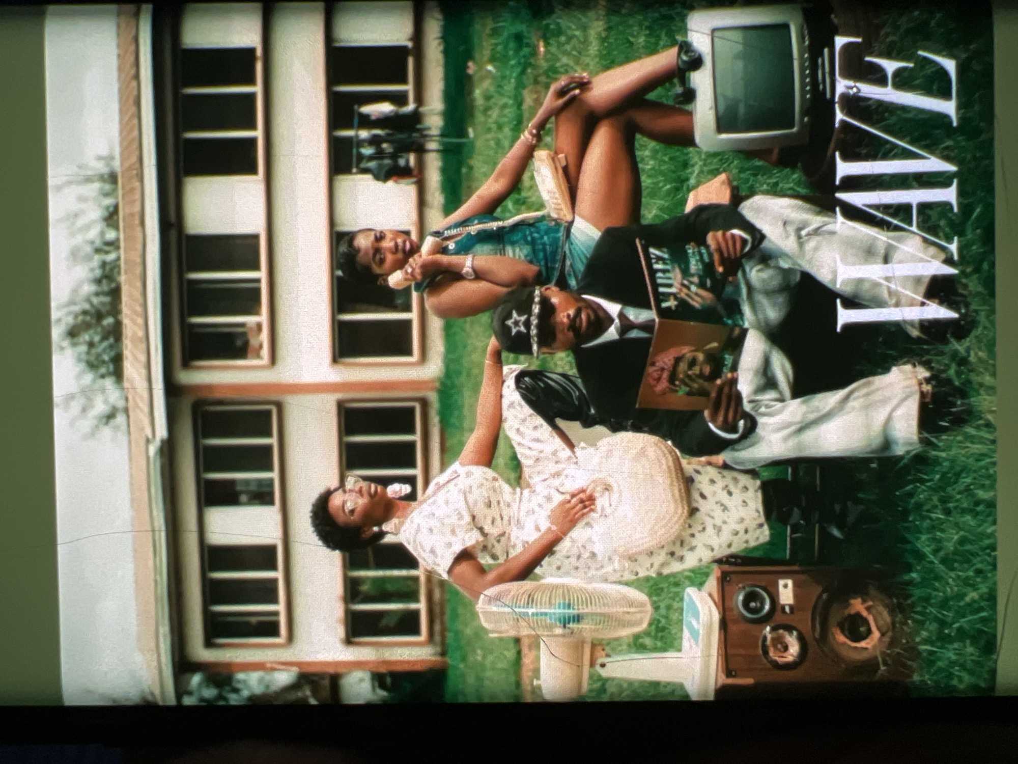 The image depicts a scene outdoors in front of a two-story building. Three individuals are present, two women and one man, all appearing to be of African descent. The woman on the left is wearing a floral print dress and glasses, holding a woven bag. To her right, a second woman is on a telephone, wearing a denim top and shorts, with jewelry on her wrist. In the foreground, a man wearing a cap, suit jacket, and tie is seated, reading a magazine titled "VIBEZ." A vintage fan, speaker, and television set are placed around the individuals. The setting is grassy, with trees visible in the background. The overall aesthetic suggests a retro or vintage theme. The architecture of the building, specifically the style of the windows and the reddish-brown roofline, points to a specific era and potentially a location with such architectural characteristics, which can be commonly found in certain districts of Lagos, Nigeria, particularly older residential or institutional buildings.