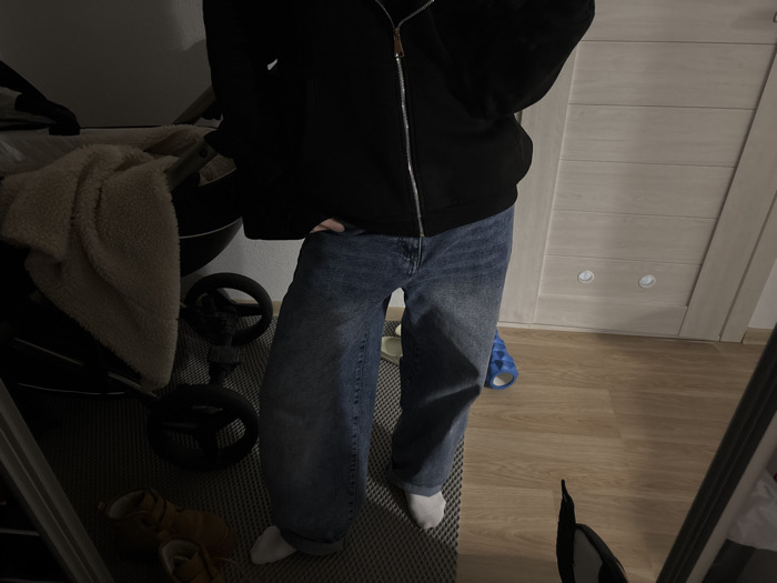 An indoor scene depicts an individual standing, partially visible from the torso downwards. The person wears a dark zip-up hoodie, blue denim jeans, and white socks. A hand is partially obscured within a jean pocket. To the left, a dark-colored baby stroller with dark wheels is present, draped with a cream-colored, textured blanket. Two small, light-brown infant shoes are positioned on a grey, textured rug beneath the stroller. To the right, a light-colored wooden door with horizontal paneling and two circular, recessed elements is visible. The floor transitions from the grey rug to light-colored wooden planks, where a single blue patterned slipper rests. A mirror on the right captures a reflection of the lower extremities of the individual and a device, likely a phone, being used to capture the image. The environment is consistent with a residential interior. No specific geographical markers or contextual details are present within the image to identify the location as Yekaterinburg, Russia.