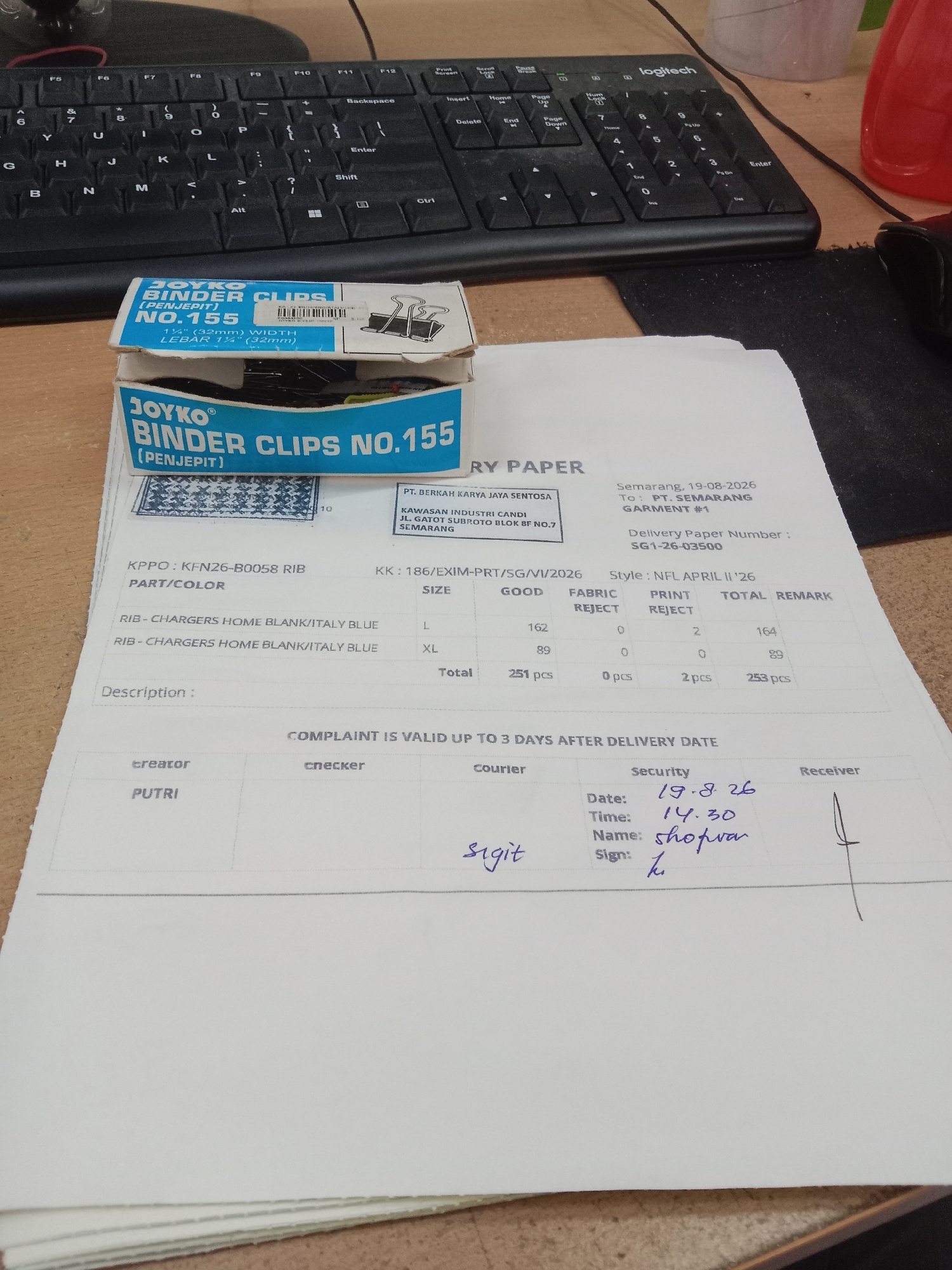 The image shows a close-up view of a desk with a keyboard and some paperwork. In the foreground, there are two open boxes of "JOYKO BINDER CLIPS NO.155". Below the boxes is a document that appears to be a delivery paper or receipt from Semarang, Indonesia, dated 19-08-2026. The document details items like "KPPO: KFN26-B0058 RIB", "PART/COLOR", "SIZE", and quantities. There are sections for "Creator", "Checker", "Courier", "Security", and "Receiver". Under the "Security" section, a date of "19.8.26", time "14.30", name "shoprvar", and signature are visible. A faint handwritten signature also appears in the "Courier" section. The background shows a Logitech keyboard. The overall environment suggests an office or workspace.