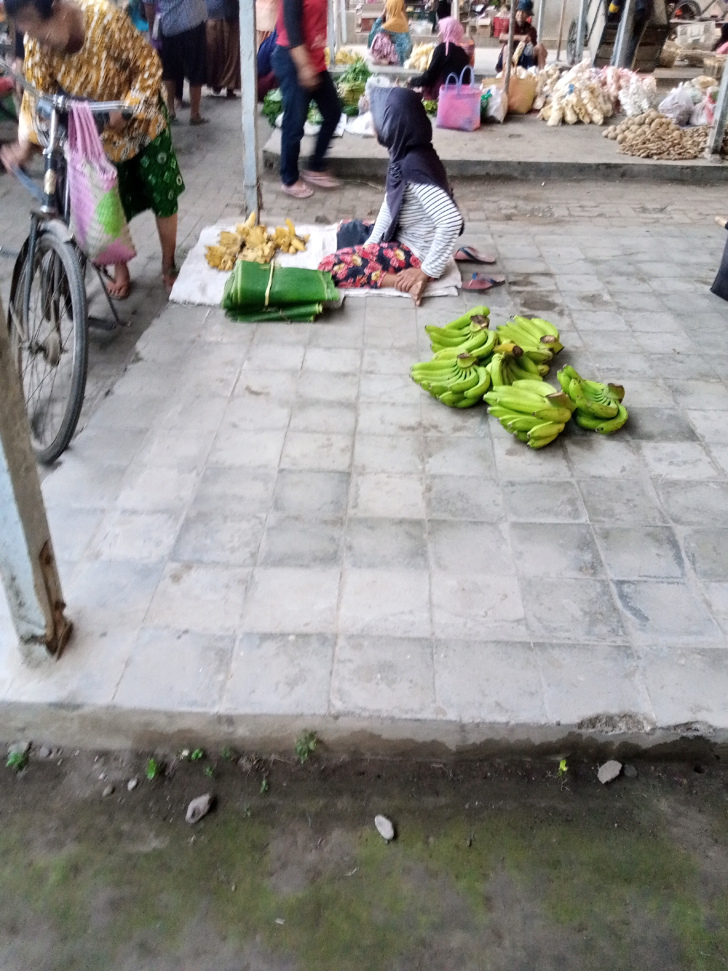 This image depicts an outdoor market scene in Ngronggot, Indonesia, likely a street market given the tiled ground and the presence of vendors. In the foreground, a woman wearing a patterned yellow and brown shirt and a green patterned skirt is standing next to a bicycle. She is leaning over the bicycle, possibly attending to it or a bag hanging from it. Her left hand is visible. On the ground, several bunches of green bananas are artfully arranged, with some stacks of green leaves bundled together.
Further back, a woman wearing a black headscarf and a striped long-sleeved shirt is sitting on a mat. She is looking downwards. Beside her are a few more bunches of green bananas. In the background, other market-goers are visible, some tending to stalls laden with produce, including what appear to be garlic or onions and potatoes. The environment is utilitarian, with a focus on the sale of goods. The ground is paved with square tiles, and there is a concrete curb separating the paved area from a patch of dirt and sparse vegetation in the lower part of the image. The overall impression is one of daily commerce in a local Indonesian market.