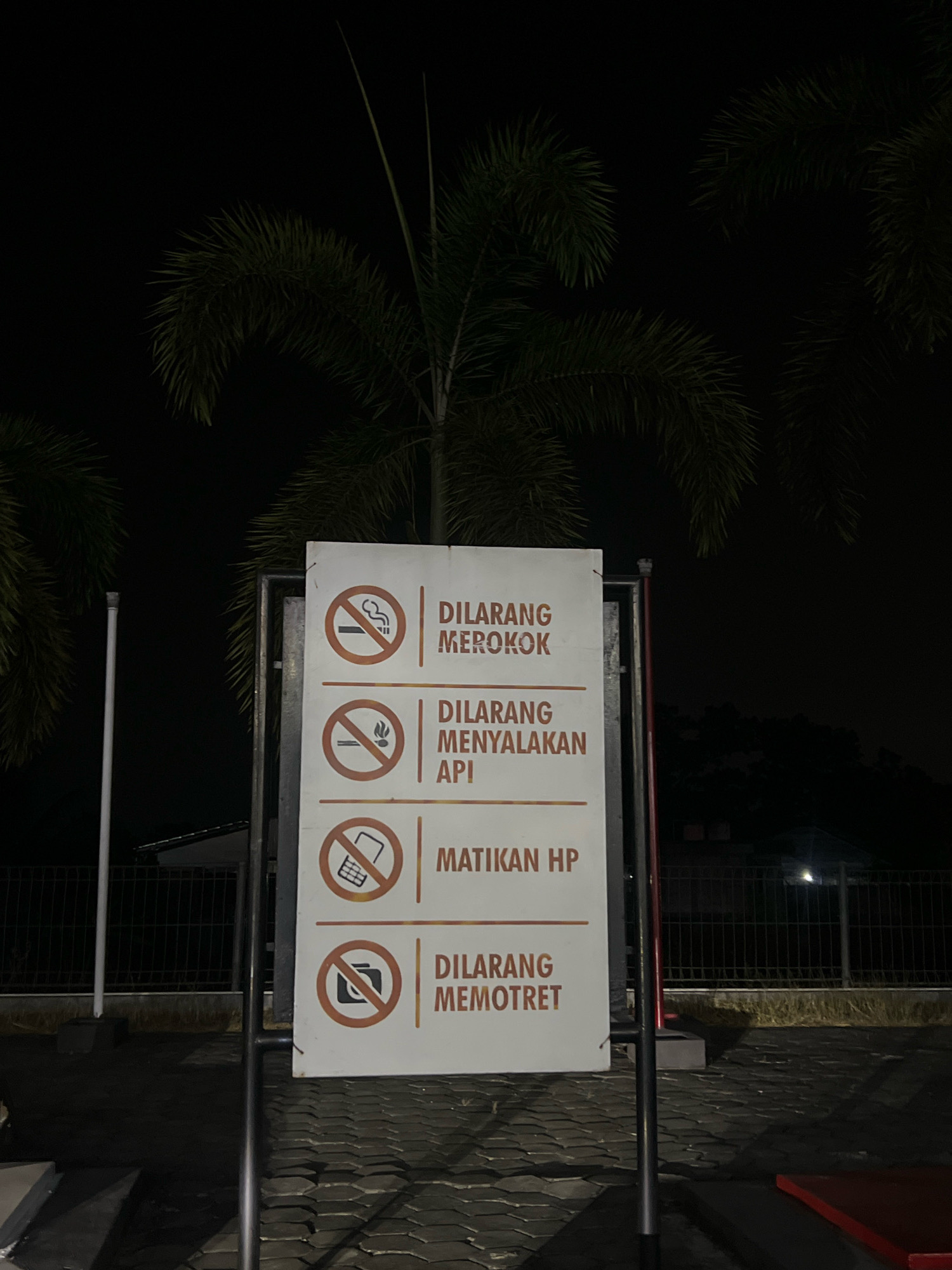 A sign in Indonesian prohibits smoking, lighting fires, using mobile phones, and taking photos. The sign is located outdoors at night, in front of a fence and some palm trees. The ground is paved with interlocking stones. The overall scene is dimly lit.