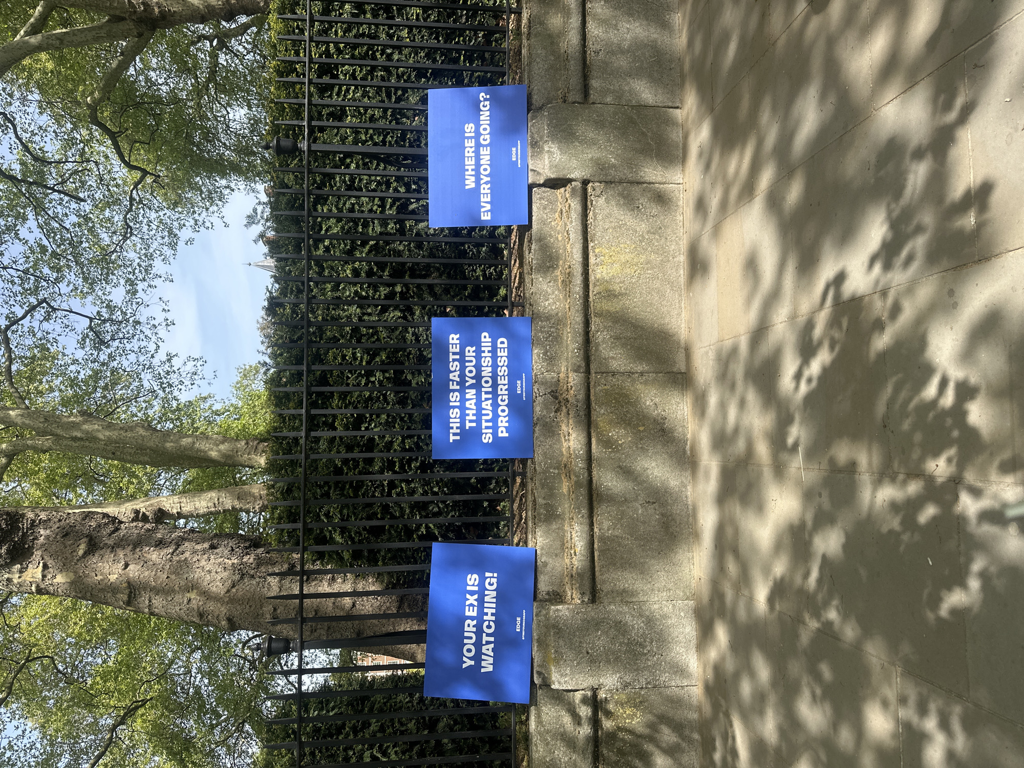 The image shows three blue signs attached to a stone wall with a black iron fence running along it. The signs, from bottom to top, read: "YOUR EX IS WATCHING!", "THIS IS FASTER THAN YOUR SITUATIONSHIP PROGRESSED", and "WHERE IS EVERYONE GOING?". Each sign also has the word "EDGE" printed on it. A tree trunk and branches are visible to the left of the signs, reaching into the sky, which is partially visible. The shadow of leaves is cast on the stone sidewalk next to the wall. The location is likely in the city of London, United Kingdom.
