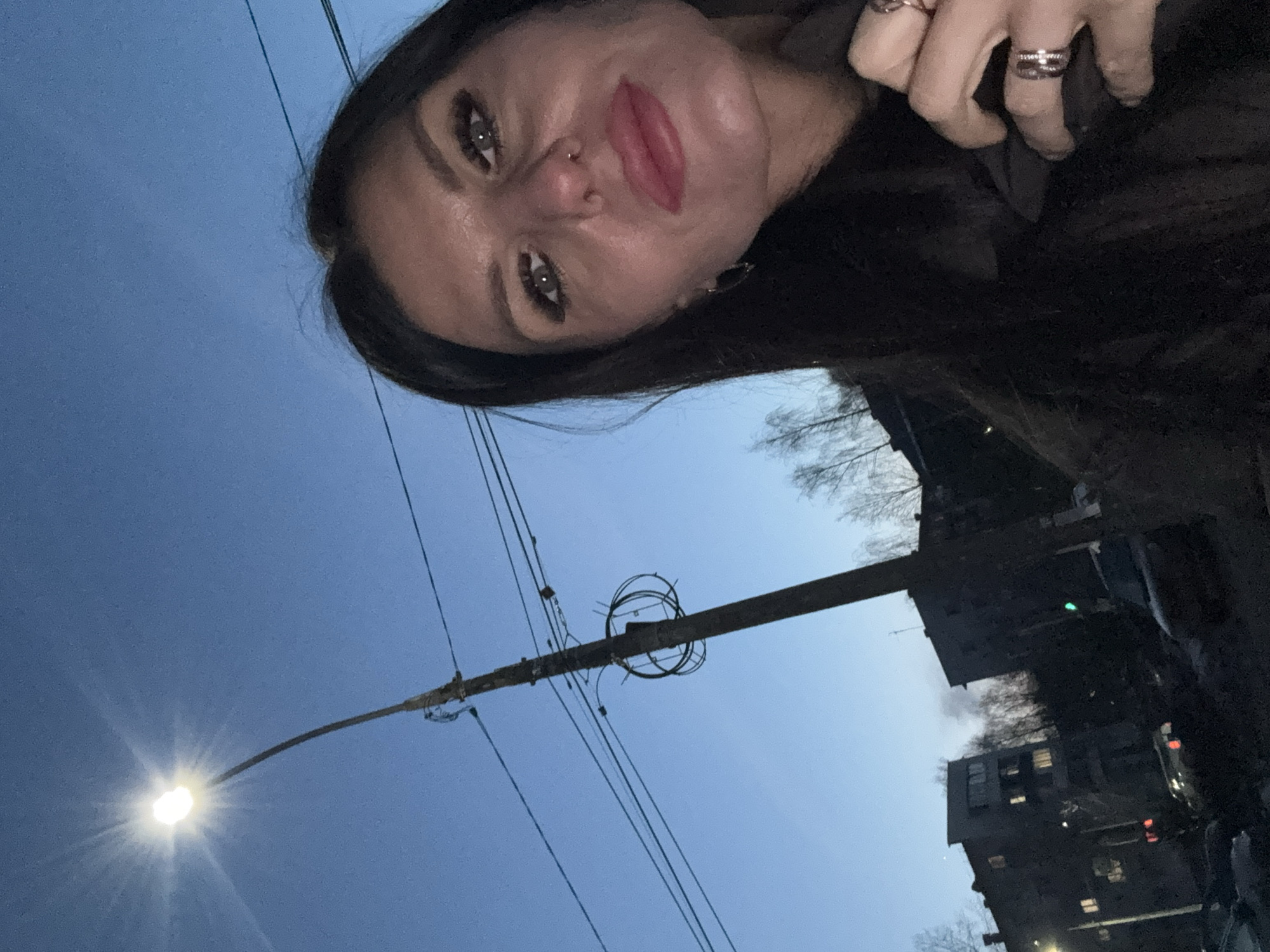 The image is a selfie taken from a low angle at dusk. The subject is a woman with dark hair and fair skin. She has a nose piercing and is wearing rings on her fingers. Behind the woman, there is a street with power lines, a streetlight, and buildings. Based on the architecture, it is likely a residential area in the city of Chaikovsky, Russia. There are a few trees visible in the background.