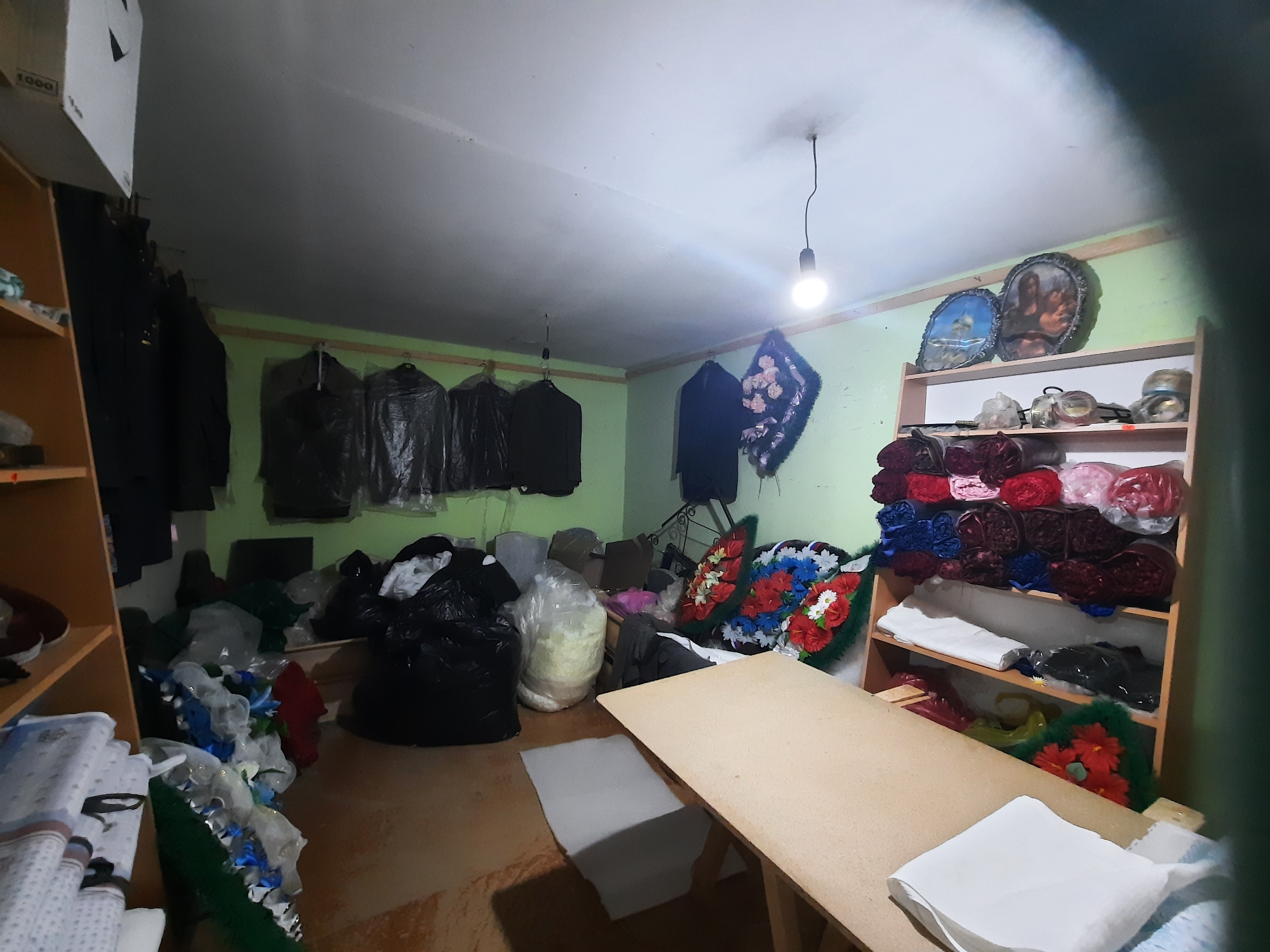 The image depicts the interior of a workshop, possibly located in Krasnogorskoye, Russia. The room is cluttered with materials and supplies. On the left, there is a wooden shelving unit filled with fabrics and other items. Garments in garment bags hang along the back wall, and two large black bags and a white plastic bag containing light-colored material are on the floor. A table occupies the right side of the frame. Stacks of white material are piled on the table. In the background is a shelf with various objects and materials. The overall impression is that of a work space that is actively used for making funeral supplies.