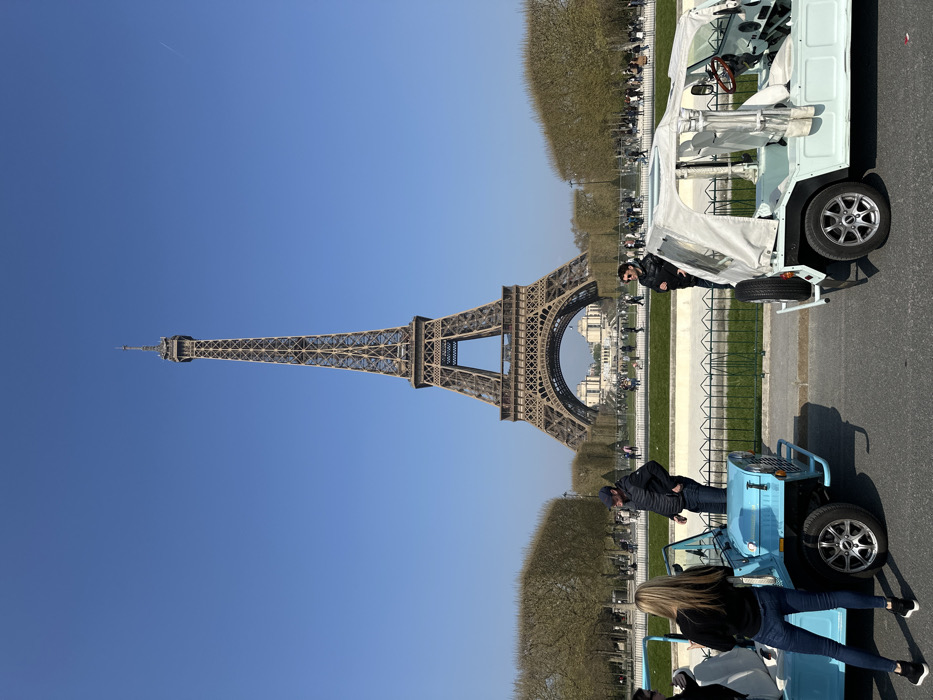 The image depicts the Eiffel Tower, located in Paris, France, viewed from the Champ de Mars looking south. The tower is centrally positioned under a clear blue sky, with the École Militaire discernible through its base in the far distance. In the foreground, two small, open-top utility vehicles, consistent with Moke-style cars, are parked on a paved surface. The vehicle on the left is light blue, and the one on the right is a light mint green with an off-white canvas roof.
Three individuals are present in the foreground near these vehicles. A woman with long blonde hair, wearing a dark long-sleeved top, blue jeans, and dark sneakers, stands with her back to the camera next to the light blue car. A man in a dark jacket, blue jeans, and a dark baseball cap stands next to the driver's side of the light blue car, facing right. Another man, wearing a dark jacket and sunglasses, stands beside the mint green car, facing left.
A low black metal fence separates the paved area from a expansive green lawn, flanked by numerous bare deciduous trees, indicating a cooler season. Numerous pedestrians are visible walking across the park grounds and around the base of the Eiffel Tower in the middle ground.