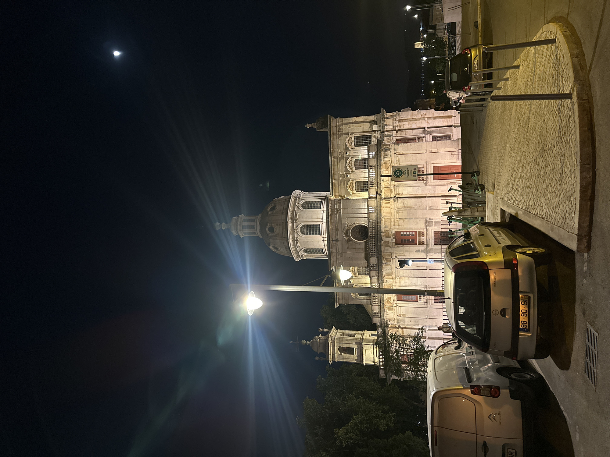 A nighttime exterior shot captures the National Pantheon in Lisbon, Portugal, illuminated by streetlights and the moon. The grand baroque building features a prominent dome, two bell towers, and numerous windows, with warm light emanating from within. In the foreground, two cars and several electric scooters are parked on a cobblestone surface. A streetlight casts a bright glare across the scene. The sky is dark, with a sliver of moon visible.