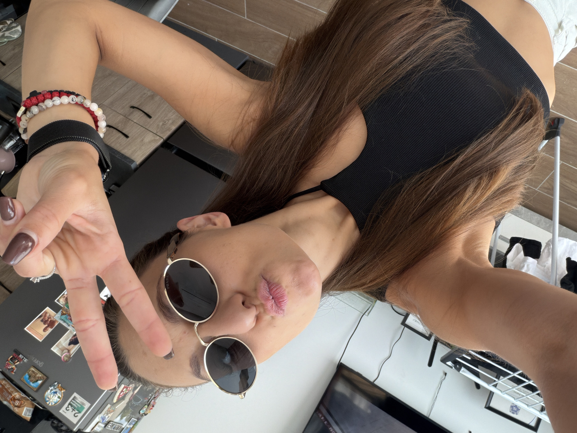 A young woman with long brown hair, wearing a black crop top and oval sunglasses, is taking a selfie. She is making a peace sign with her right hand and puckering her lips. She has on several bracelets on her left arm and a ring on her right ring finger. In the background, there is a refrigerator with magnets and photos, as well as some furniture and a television screen. The setting appears to be an indoor apartment, likely in the city of Plovdiv, Bulgaria.