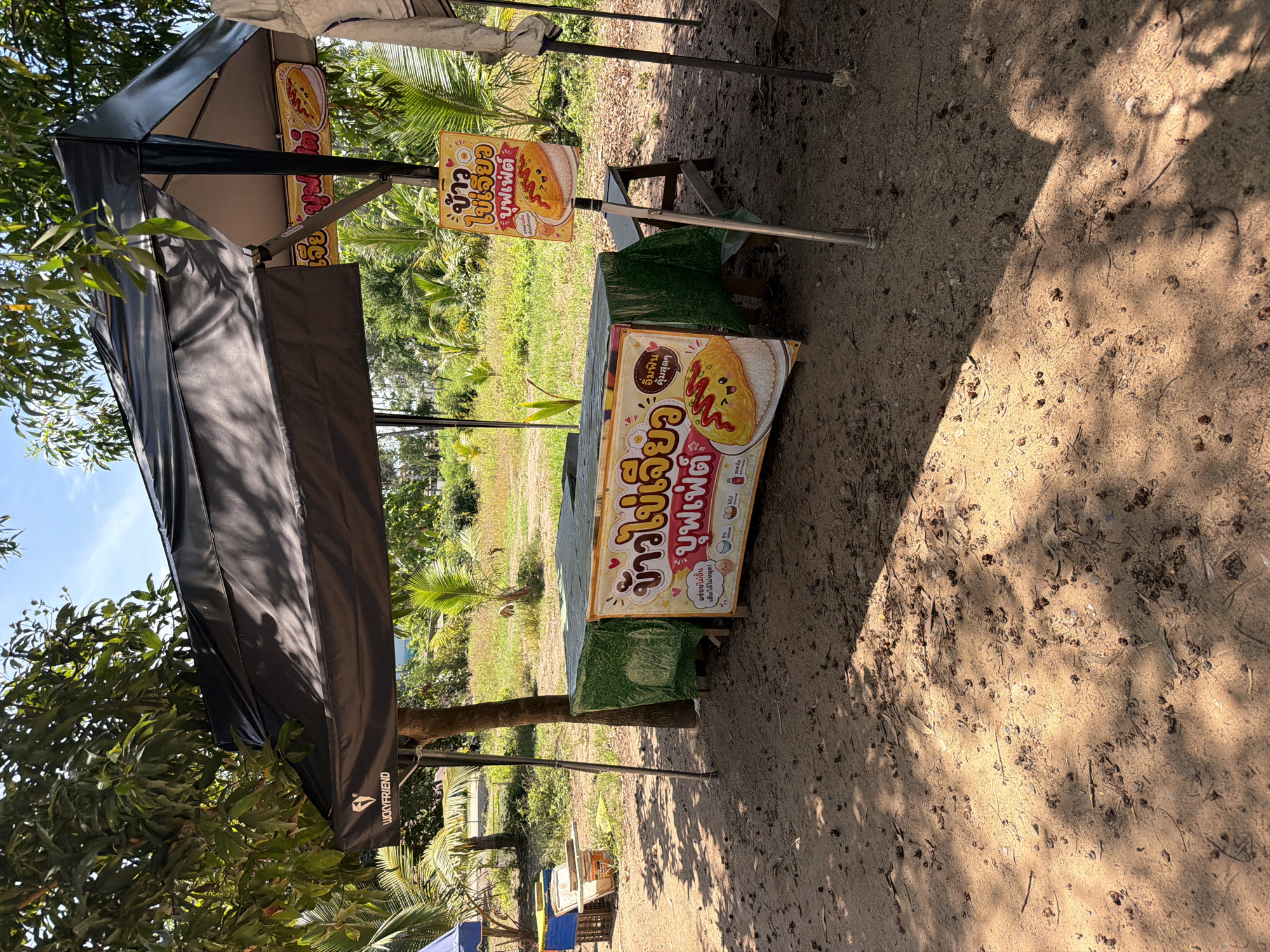 The image shows an outdoor food stall in Baan Bo Phut, Thailand, situated on a sandy area under the shade of trees and a black canopy. The stall is set up with a green-covered table displaying food items. Two signs are visible, one attached to the table and another mounted on a pole. The sign on the table features a cartoon image of an omelet with a smiling face and ketchup drizzle, accompanied by Thai text and smaller images of food. The sign on the pole also depicts a similar omelet and has Thai text. In the background, there is a lush green landscape with palm trees and other vegetation, under a clear blue sky. To the left of the stall, some blue and yellow crates are stacked. The overall scene suggests a casual, open-air food vendor operating in a tropical environment. No people are visible in the image.