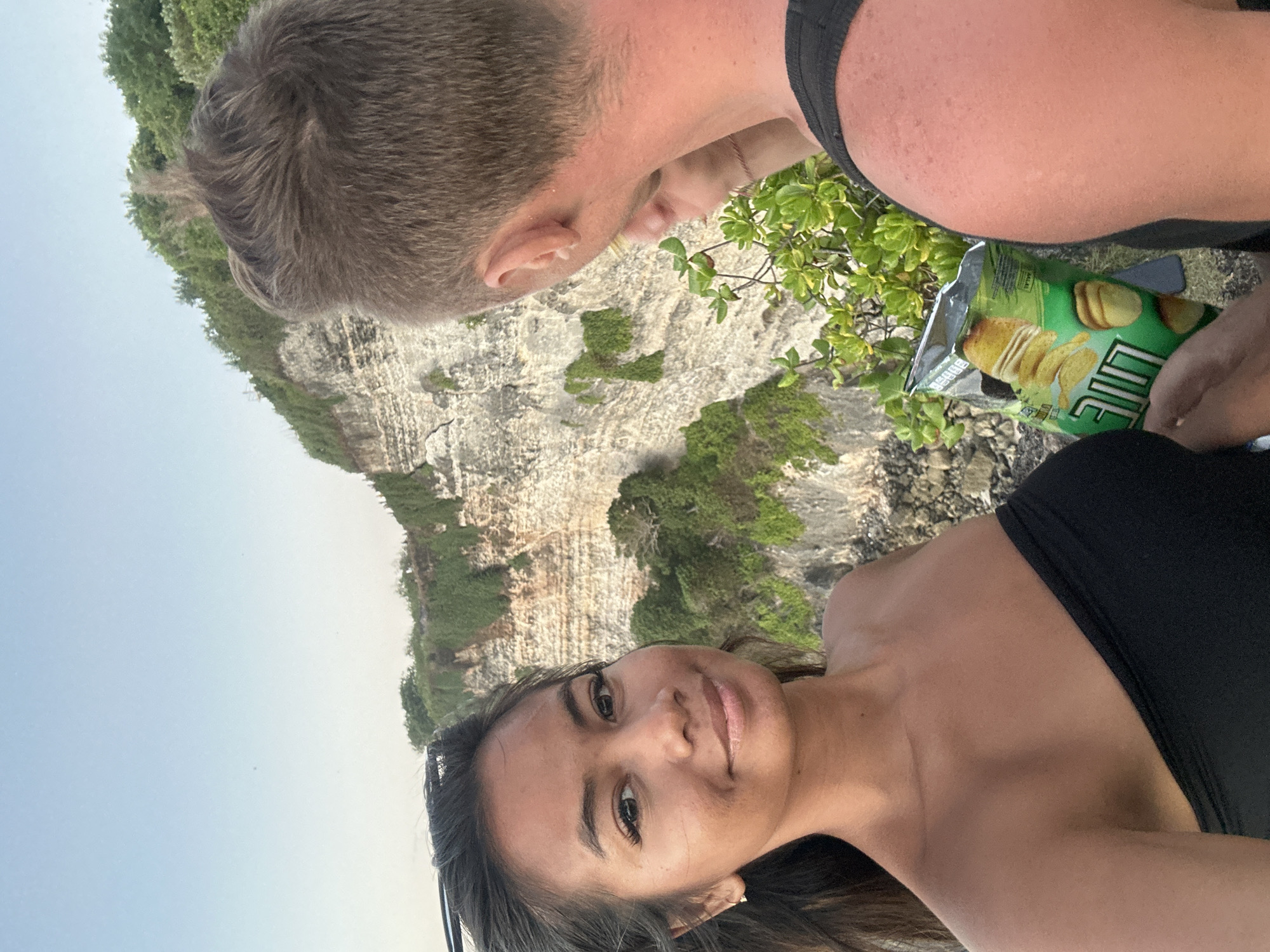 In the city of Kuta Selatan, Indonesia, a woman with dark hair is captured in a close-up selfie, smiling at the camera. She is wearing a black strapless top. To her right, a man with short, spiky brown hair is seen from the back, looking over his shoulder towards the woman. A partially opened bag of chips, with a green and yellow design, is held by someone off-camera in the lower right portion of the frame. The background reveals a dramatic cliff face covered in lush green vegetation, with a clear blue sky above. The rough texture of the cliff and the dense foliage suggest a natural, rugged landscape. The lighting indicates it is either sunrise or sunset, casting a soft glow over the scene.