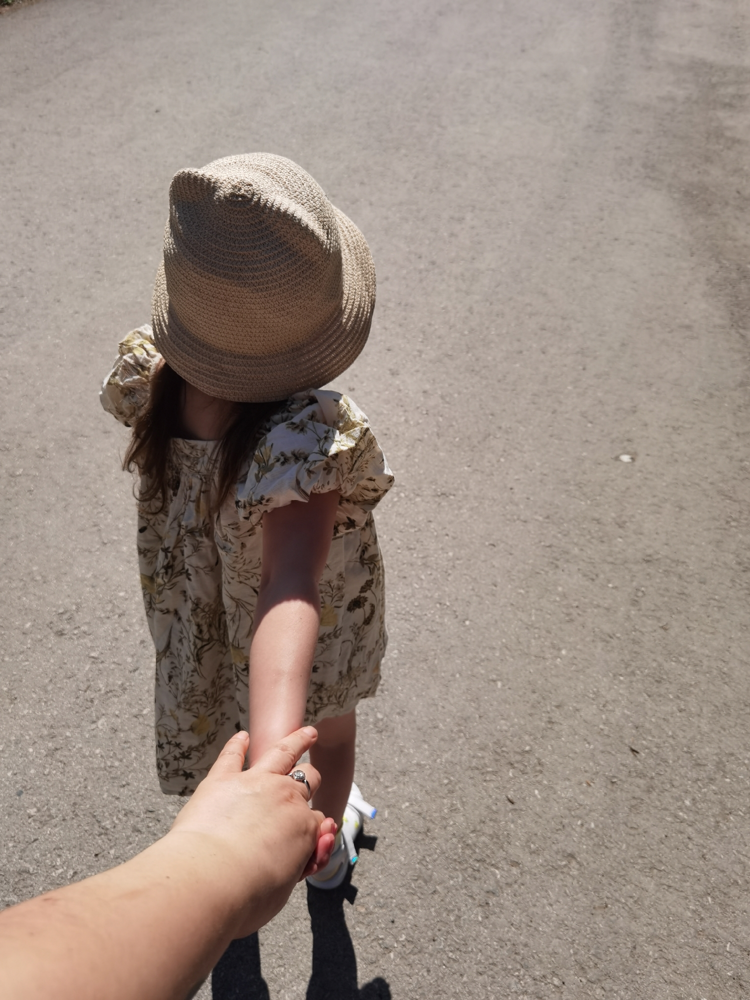 A young child in a floral dress and straw hat is being led by the hand on a paved surface, likely a street or sidewalk. The child's back is to the viewer, obscuring the face beneath the hat. An adult's hand, adorned with a ring, is holding the child's hand. The paved surface is gray and shows signs of wear. The location is unknown.