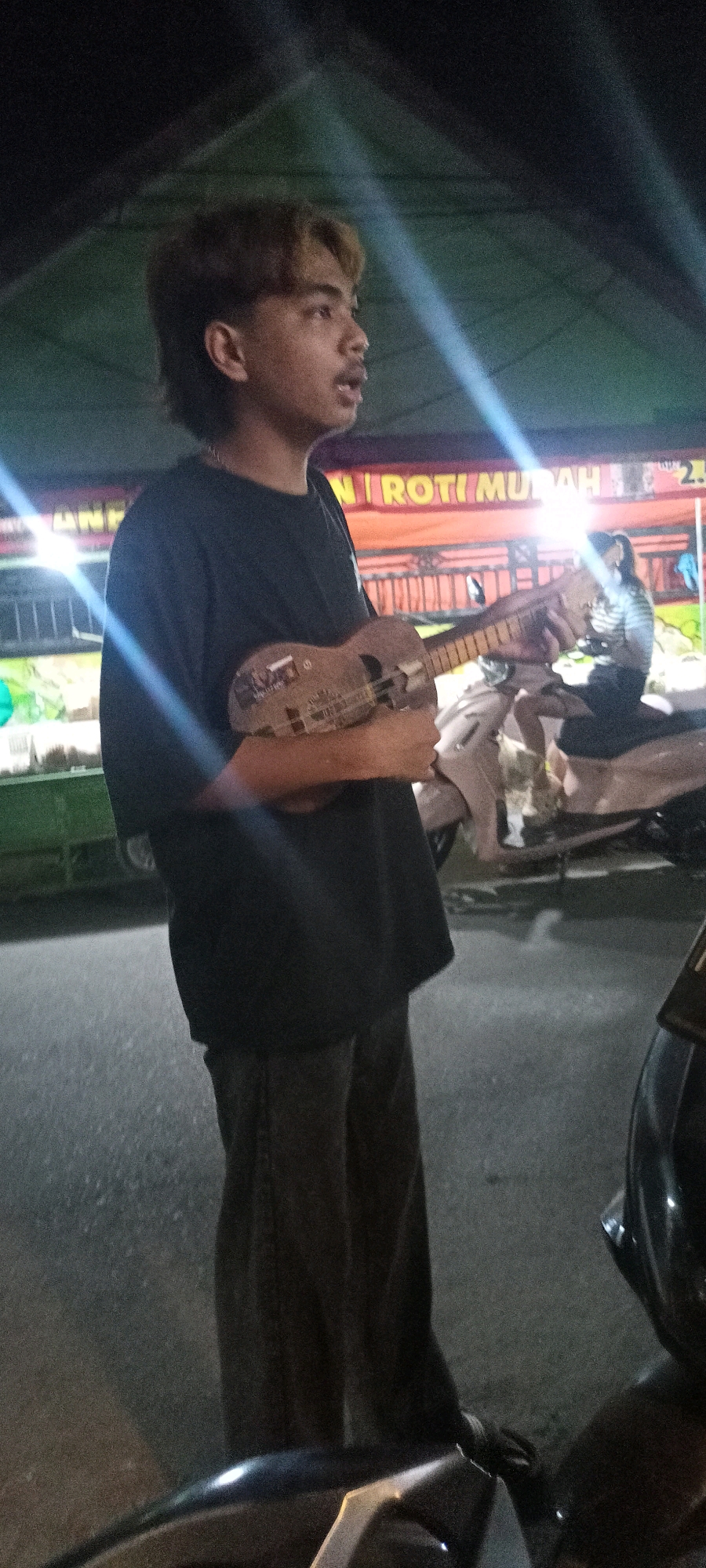 A young person with dyed hair, wearing a black t-shirt and dark pants, stands in the foreground holding a ukulele. The person is looking to the right, with their mouth slightly open. Behind them, a person sits on a motorcycle. The background features a tent with orange fabric displaying Indonesian text, likely a food stall, with the words "ROTI MUDAH" (Easy Bread) visible. Bright lights streak across the image, suggesting an outdoor nighttime setting. The ground is asphalt. The overall scene appears to be a street in Bendotretek, Indonesia.