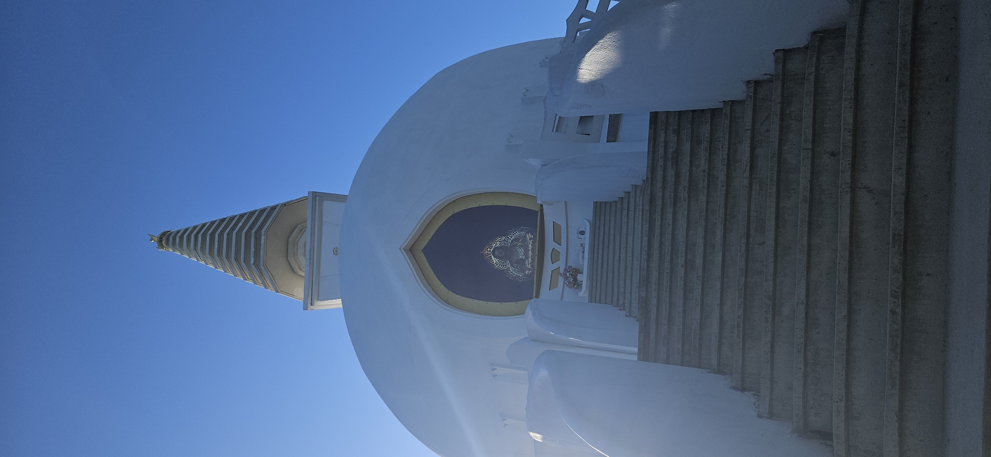 The image shows a white stupa with a tiered spire, set against a clear blue sky. A statue of Buddha is visible within an arched recess at the base of the stupa. A set of concrete stairs leads up to the recess. The stupa is the Béke-sztúpa, or Peace Stupa, located in Zalaszántó, Hungary. The scene is observed from a low angle looking upwards. The lighting suggests it is daytime, with sunlight casting shadows on the stupa. No people are present in the image.