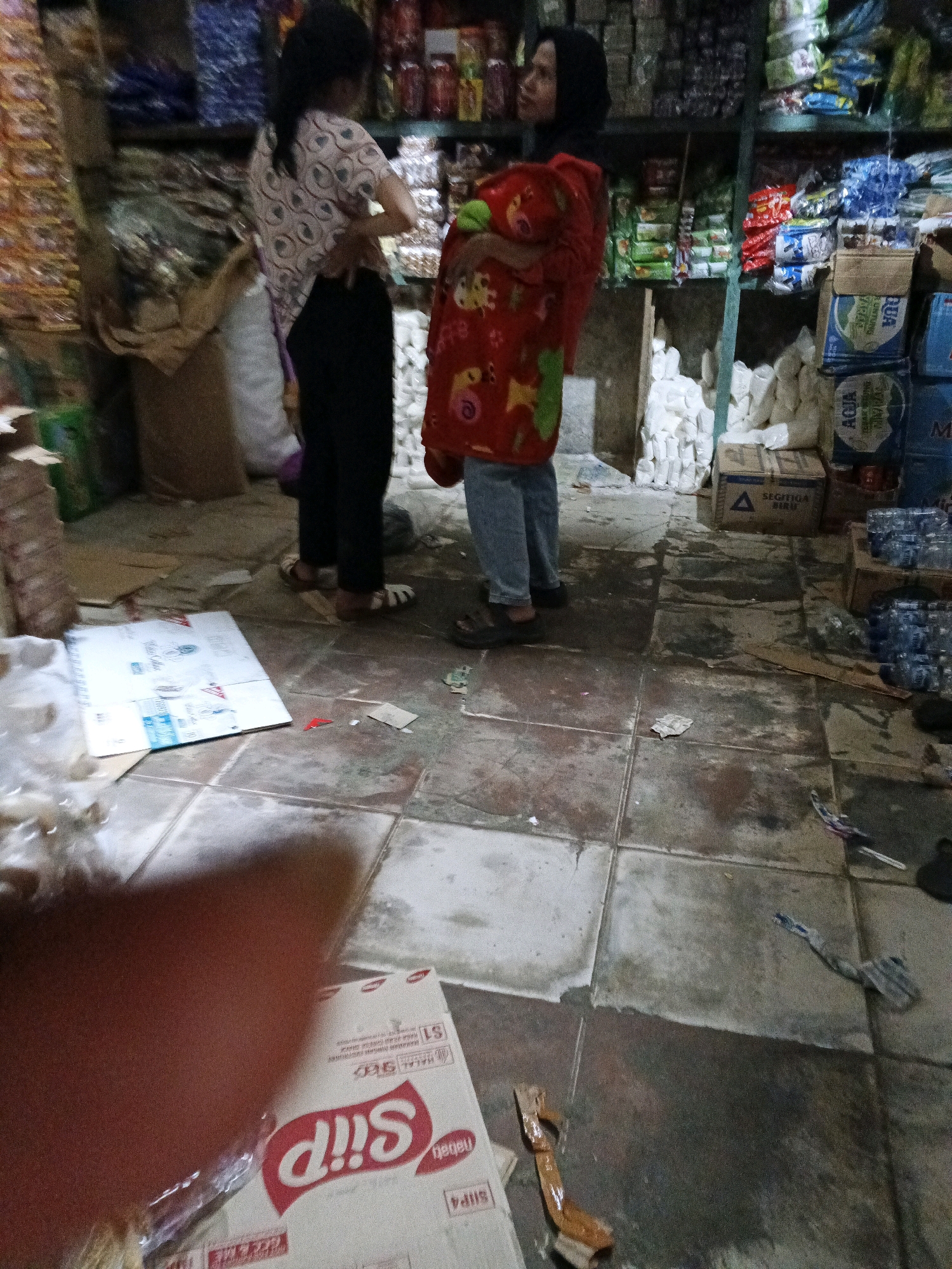 The image shows an indoor scene, likely a shop or storage area, in Warungkiara, Indonesia. Two women are present. One woman, on the left, wears a patterned shirt and dark pants, and is facing away from the camera with her hand on her hip. The second woman, on the right, wears a black headscarf and a red blanket over a blue shirt and jeans. She is also facing away from the camera, and appears to be holding something green. The floor is tiled and appears wet in places. Shelves are visible in the background, filled with various packaged goods. A large cardboard box with "Sip" branding is visible on the floor in the foreground, along with other scattered items.