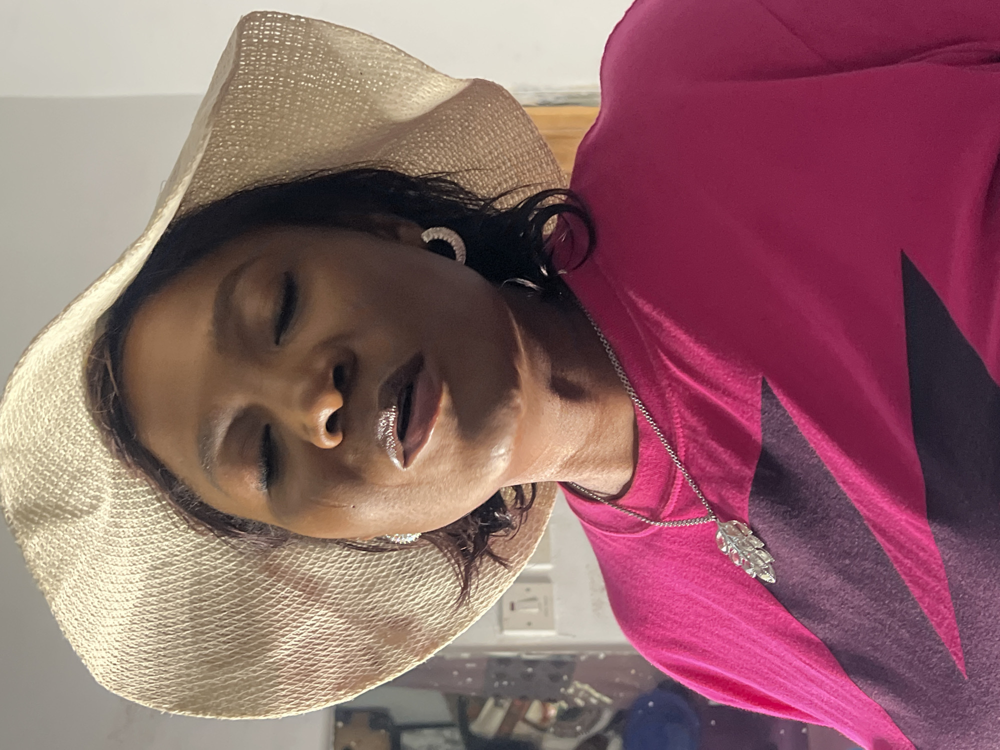 A close-up, medium shot shows a Black woman wearing a wide-brimmed straw hat and a vibrant pink long-sleeved shirt with a purple geometric design. She has her eyes closed and her mouth slightly open, with her lips glossed. She is wearing hoop earrings and a silver pendant necklace shaped like a feather. The background is out of focus, with hints of a domestic environment, possibly a bedroom, with what appears to be a light switch on the wall and some shelving units. The lighting suggests an indoor setting, possibly during the day. The woman's pose and expression could imply a moment of reflection or prayer. There are no explicit indications of the location being Jos, Nigeria, in the image itself, but based on the provided context, this scene is assumed to be set in Jos, Nigeria.