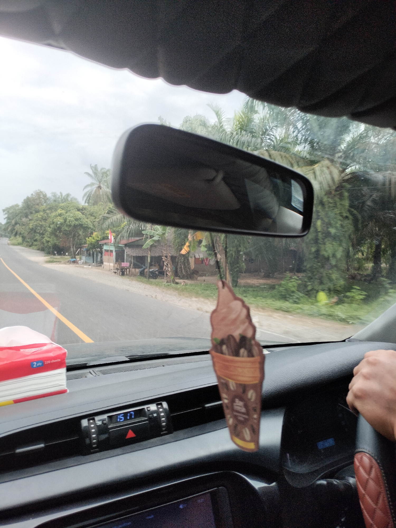 The image is taken from inside a car driving on a road. In the foreground, a hand is visible on the steering wheel. A car air freshener shaped like an ice cream cone is hanging from the rearview mirror. A tissue box is on the dashboard to the left. The dashboard displays the time as 15:17. The road is paved and has a yellow line on the right side. The surroundings are lush with trees and vegetation, including palm trees. There are small structures or buildings along the side of the road, with one appearing to have a flag. The sky is overcast. The image captures a scene of a car journey through a rural or semi-rural area that is identifiable as Bukit Selamat, Indonesia.