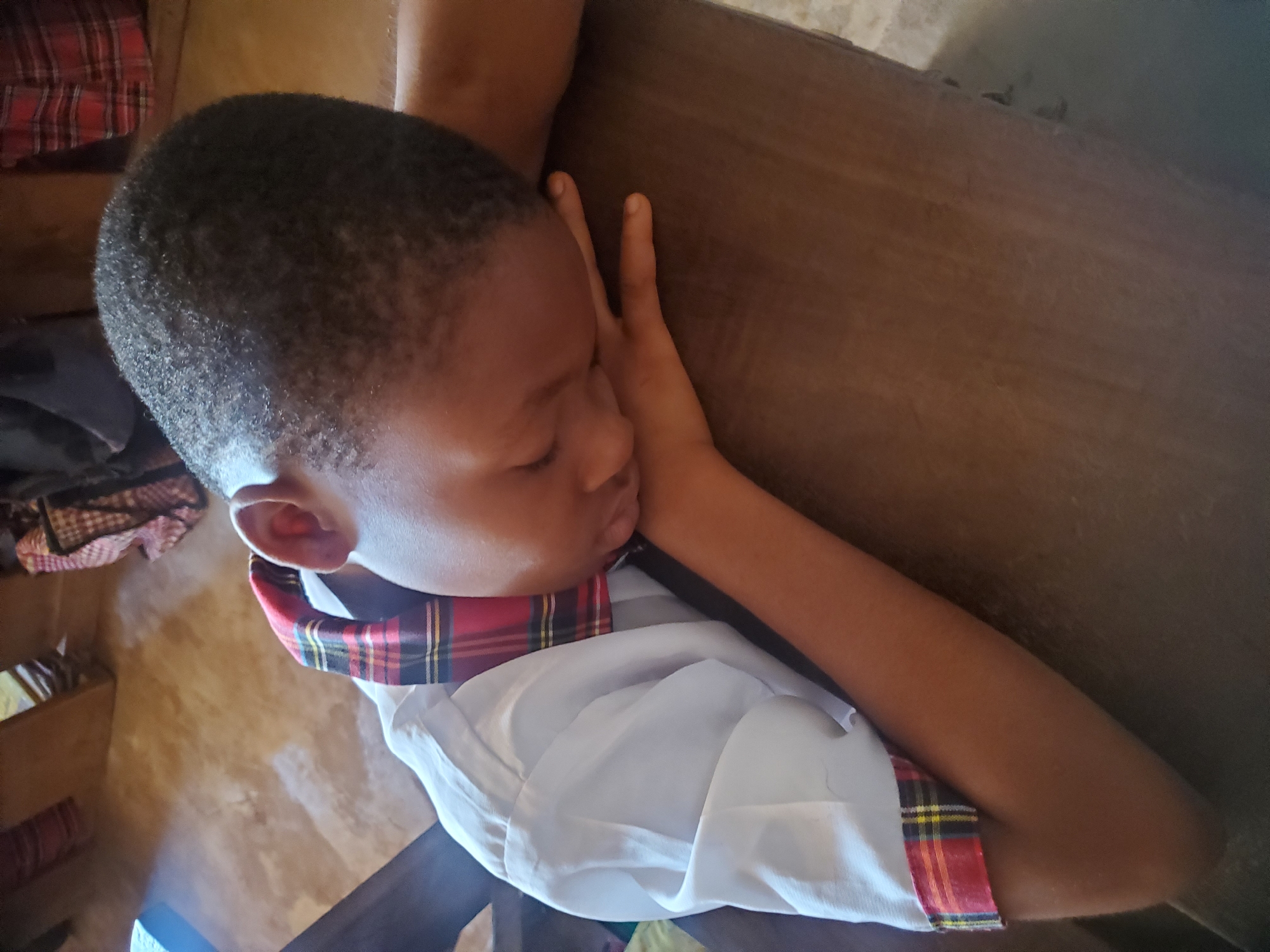 The image depicts a child, presumably male, resting on a wooden surface. The child is wearing a white and plaid uniform, and appears to be sleeping with his arm supporting his head. The background includes a glimpse of the floor and possibly other items, suggesting an interior space. The image was taken in Ukwu-Abwa, Nigeria.