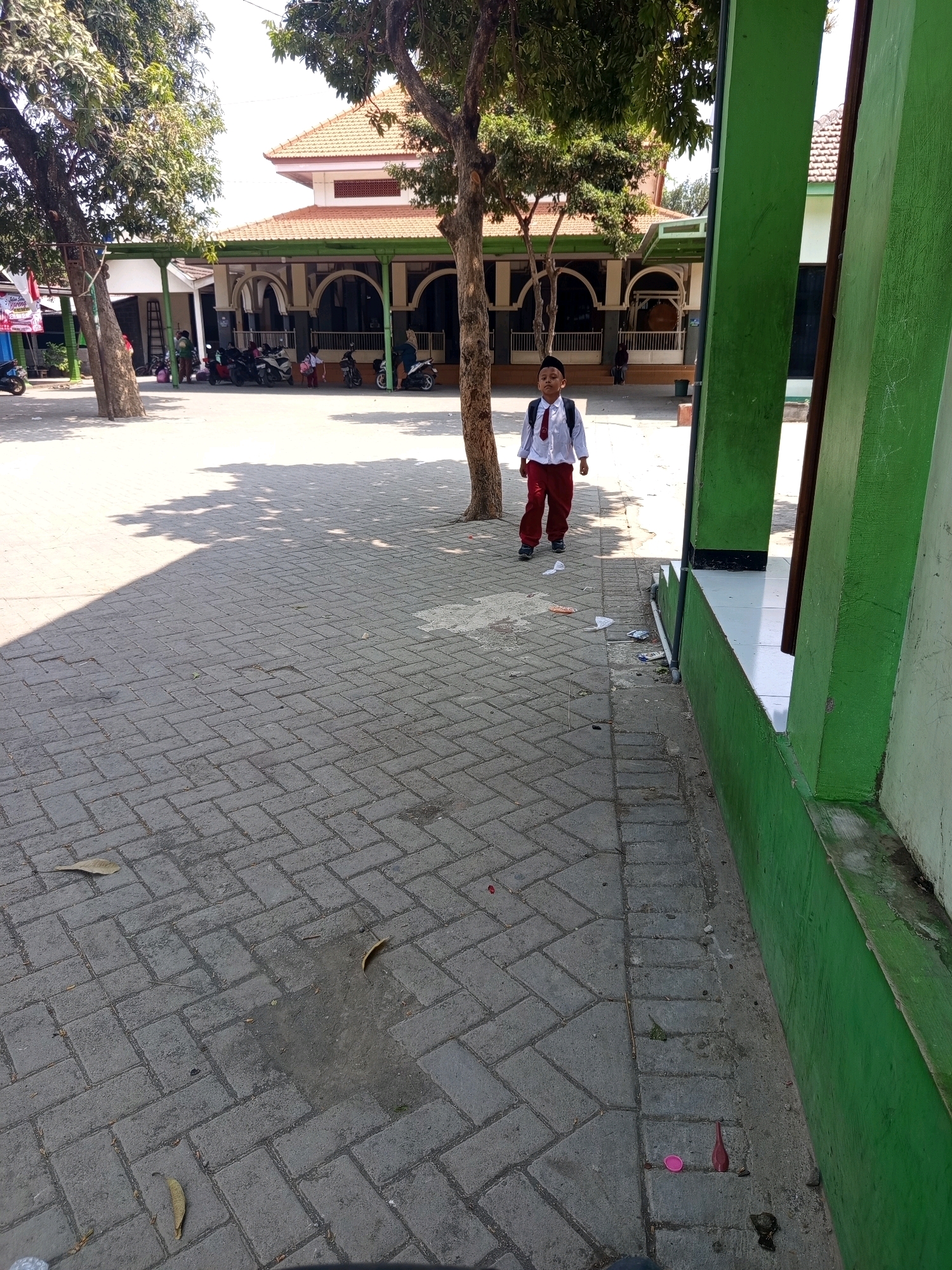 A young boy in a school uniform walks on a paved courtyard in front of a building with a red tiled roof and green accents. The boy is wearing a white shirt, a red tie, and red pants, and he has a backpack on his back. The courtyard is made of grey interlocking pavers and is partially shaded by trees. In the background, there are several motorcycles parked. The building appears to be a school or religious institution, typical of the architecture found in Gondanglegi, Indonesia. The scene is captured during daylight hours, with bright sunlight casting shadows on the ground. The overall environment suggests a typical day at a school in a tropical climate.