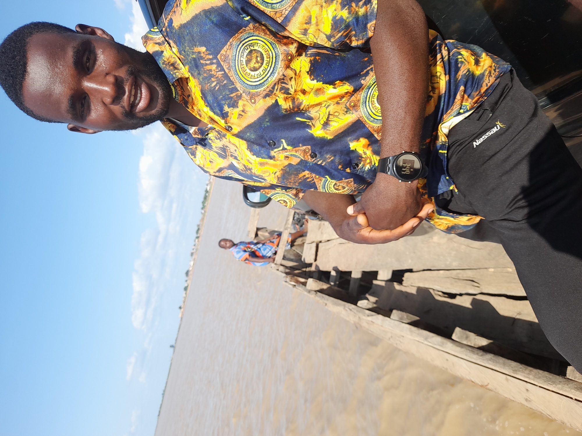 In Ibi, Nigeria, a Black man with a beard and short dark hair smiles at the camera, wearing a colorful short-sleeved shirt with a bold pattern and black pants. He has a black digital watch on his left wrist. He is leaning against a dark car, with his hands clasped in front of him. In the background, a wooden boat is partially visible, filled with muddy water. Another person, also Black, is sitting in the boat, wearing a blue and orange patterned shirt. The sky is blue with scattered white clouds, and the water appears to be a large river. The overall scene suggests a sunny day near a body of water.