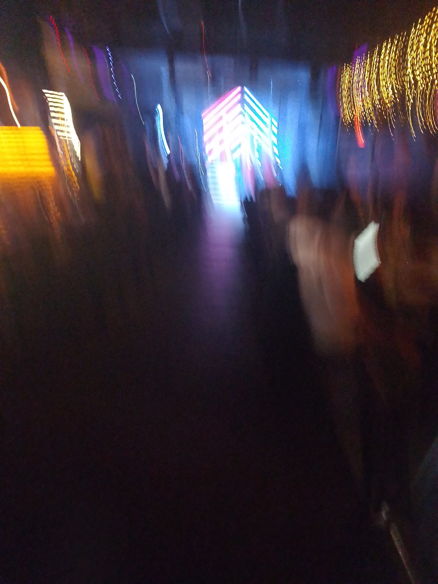 The image, taken with motion blur, appears to show a performance or event in an indoor venue, possibly in the city of Chaikovsky, Russia. A brightly lit structure or stage design is visible in the center, with blue and white lights. Yellow and orange lights are present on the left and right sides of the scene. People are blurred in the foreground and background, suggesting a crowded event. The ground is dark and undefined.