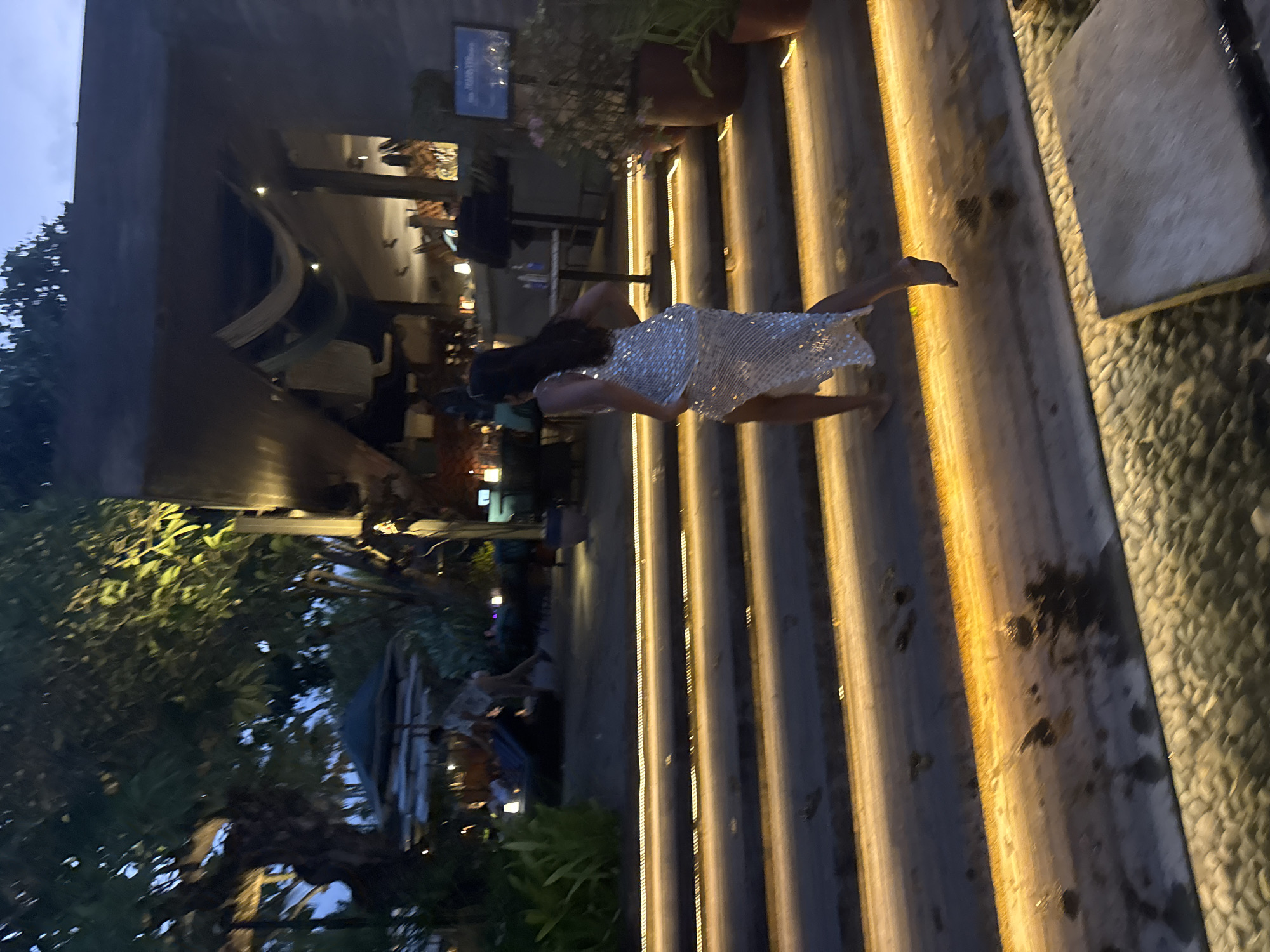 A woman in a sparkling silver dress with a high slit is running up a set of outdoor stairs at night. The stairs are illuminated by a warm, yellow light embedded in the edges of each step. The woman is barefoot and her hair is dark and long. Behind her, the scene transitions to a dimly lit outdoor dining or bar area with tables and chairs visible. There are trees with lush foliage and a dark sky overhead. In the background, there are hints of a building with an awning and some potted plants. The overall impression is of a stylish evening venue.