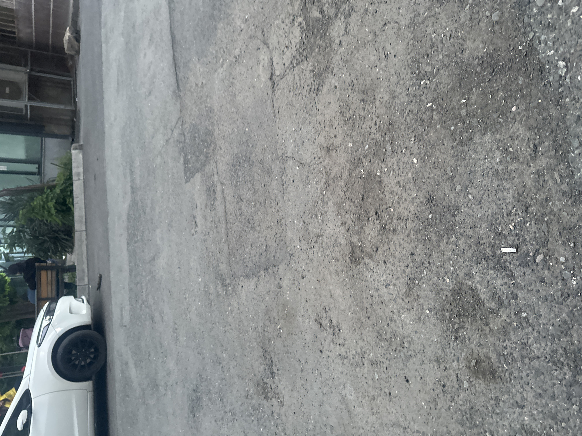 The image shows a section of a paved street in Yerevan, Armenia. The street surface is predominantly grey and shows signs of wear and tear, with a mix of smooth patches and areas with gravel. A white car is partially visible in the lower-left corner, adjacent to a building with glass windows and greenery. A cigarette butt lies on the street surface.