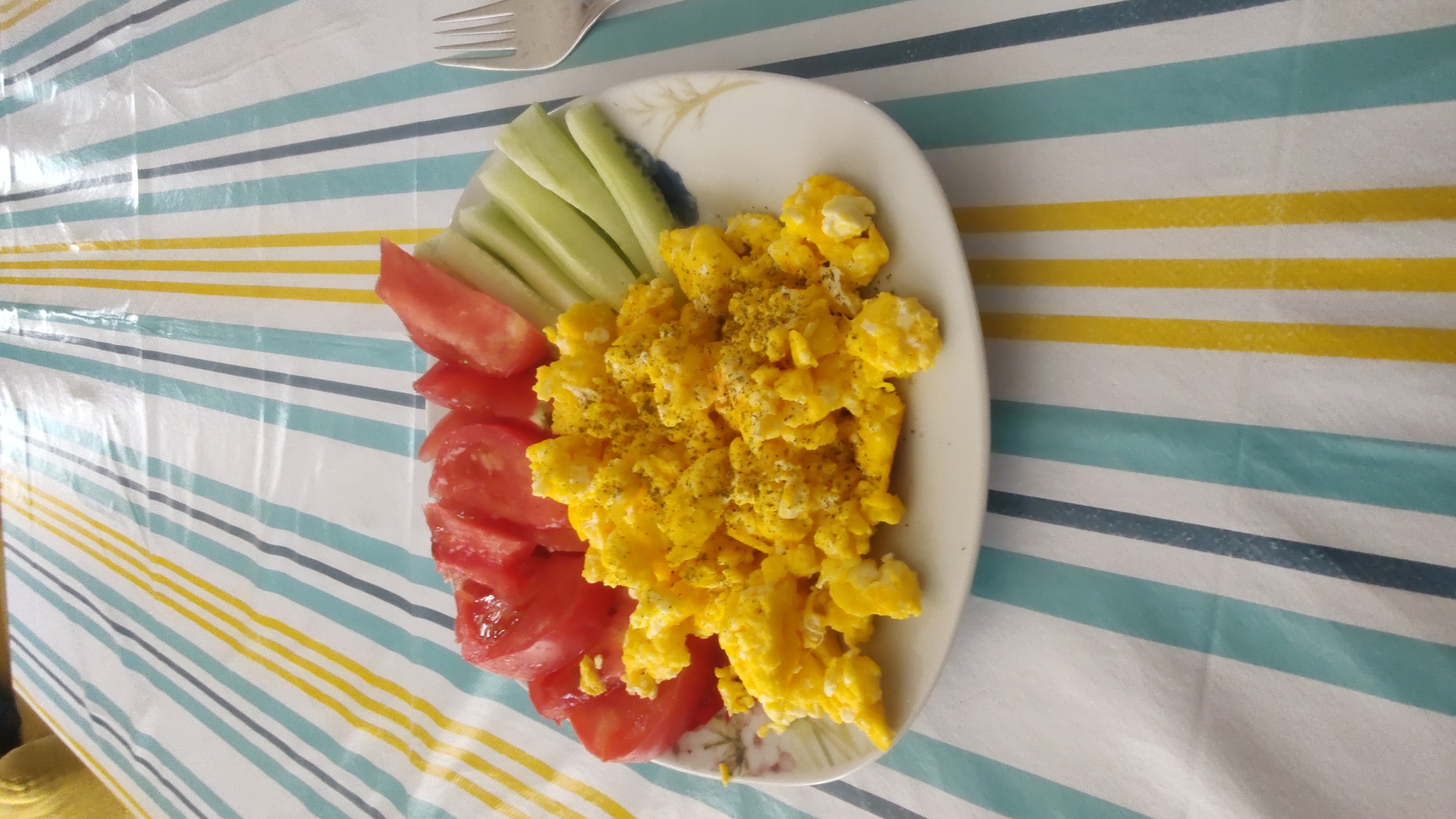 A close-up, top-down shot showcases a plate of food on a striped tablecloth. The plate contains scrambled eggs seasoned with pepper, sliced tomatoes, and cucumber sticks. The tablecloth features alternating blue, yellow, and white stripes. A silver fork is partially visible on the right side of the frame. The location is identified as Kurtovo Konare, Bulgaria.