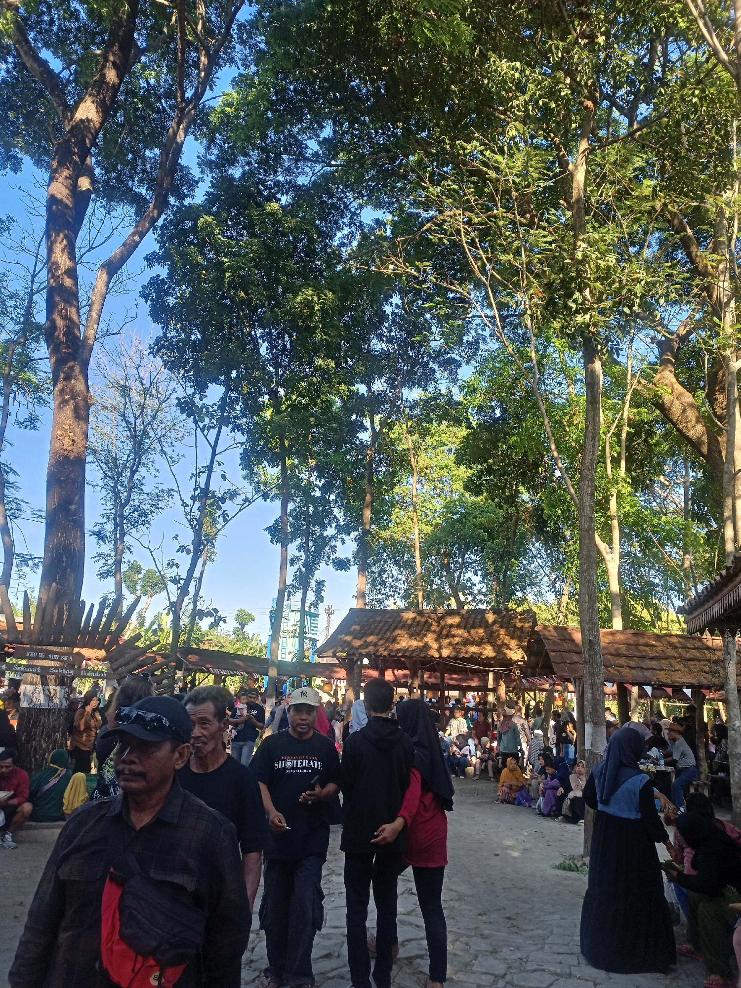 In Karungan, Indonesia, a scene unfolds beneath a canopy of large trees and a bright blue sky. The ground is paved with a rough, gray stone, and the overall atmosphere suggests a bustling outdoor market or festival. Several wooden stalls with thatched roofs are scattered throughout the area, offering shelter and housing various goods or services.
The foreground is dominated by a group of people. Closest to the viewer, a man wearing a black plaid shirt and a black baseball cap with sunglasses perched on top, is looking to his left. He has a red bag slung across his chest. Behind him, another man in a black t-shirt is walking towards the right, and a third man in a black t-shirt with the words "PERSATUAN SHOTORATE" is visible to his right.
Further into the scene, many other individuals are gathered. Some are seated, while others are standing and interacting. A woman in a red shirt and black headscarf is walking with another person in black. In the background, more stalls are visible, and a blue structure, possibly a tower or building, can be seen through the trees. Signs with Indonesian text are present on some of the wooden structures, indicating the location and potentially the nature of the event. The lighting suggests it is daytime, with sunlight filtering through the leaves and casting shadows.