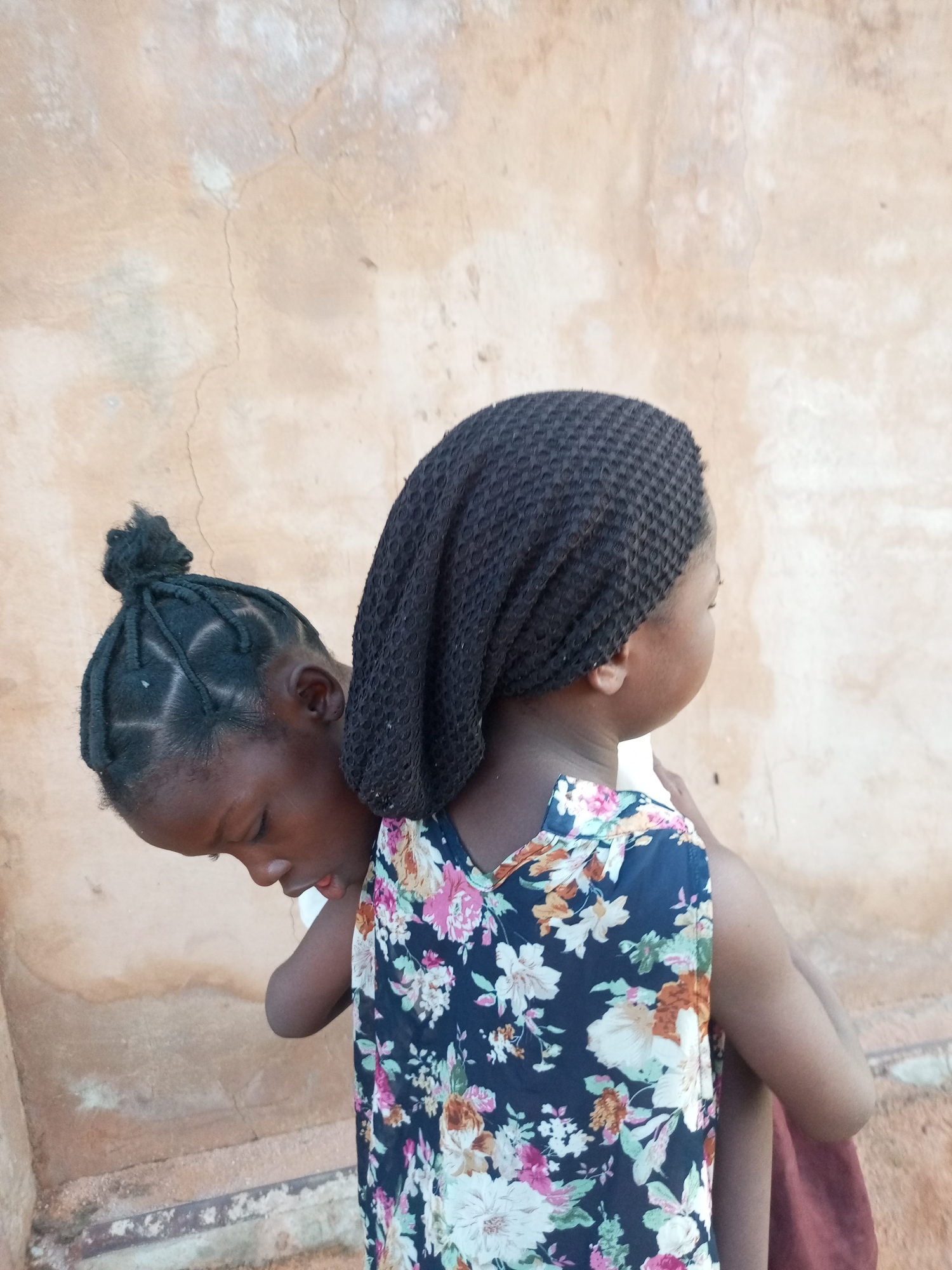 Two young Black girls are depicted from the back, standing in front of a textured, light-brown, aged wall. The girl on the left has her hair styled in braids pulled into a bun on top of her head and is leaning forward, her face partially obscured as she looks down. The girl on the right wears a dark blue sleeveless dress with a vibrant floral print featuring pink, white, and orange flowers, and a black head covering. The wall behind them is weathered, with visible cracks and patches of discoloration, suggesting an outdoor or semi-outdoor setting possibly in a residential area of Tempure, Nigeria.