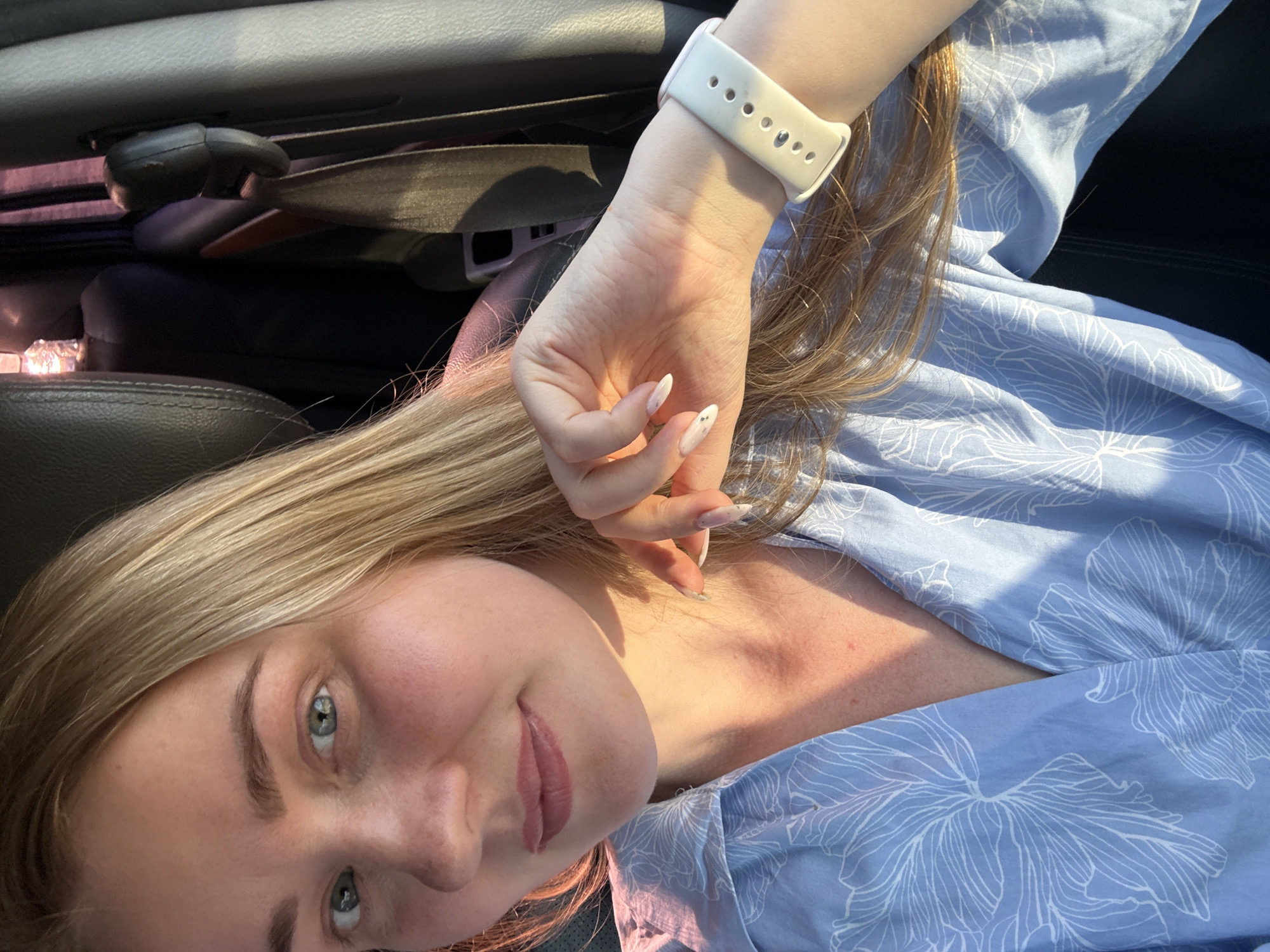 A selfie angled from above shows a fair-skinned woman with blonde hair and light eyes. She wears a light blue patterned shirt and a white smartwatch on her left wrist. The background appears to be the interior of a car, with dark-colored seats and a seatbelt mechanism visible. The location is not specified, but the request includes the city of Chaikovsky, Russia, though there is no information in the image to support this.