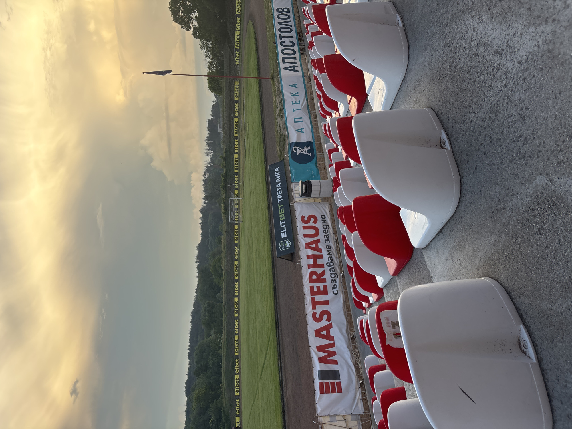 The image displays an empty sports stadium, likely a football venue in Kazanlak, Bulgaria, captured during the late afternoon or early evening. The scene is viewed from the spectator stands, looking across the pitch towards a tree-lined horizon under a striking sky.
The sky, occupying the upper left portion of the image, is filled with dramatic clouds illuminated by warm, golden light, indicative of either a sunrise or sunset. In the foreground and extending to the right are rows of clean, empty stadium seats, alternately colored red and white. These seats are attached to concrete risers.
The middle ground features a well-maintained green grass football pitch, surrounded by a dirt track often found in multi-purpose stadiums. Along the perimeter of the pitch, numerous advertising banners are visible. Repeated text "efbet" points to a prominent sponsor. Other banners clearly display "ELITEBET ТРЕТА ЛИГА," identifying a professional football league. Additional advertisements include "АПТЕКА АПОСТОЛОВ" (Apostolov Pharmacy) and "MASTERHAUS създаваме заедно" (Masterhaus - we create together). A partial inscription "P03A," likely "РОЗА" (Roza - Rose) in Bulgarian, can be seen on one of the white seats. A tall, dark, pointed flag or banner stands out against the bright sky.
The stadium appears quiet and unoccupied, suggesting a moment between events or outside of game hours. The distant treeline and the dynamic sky provide a serene backdrop to the man-made structure.